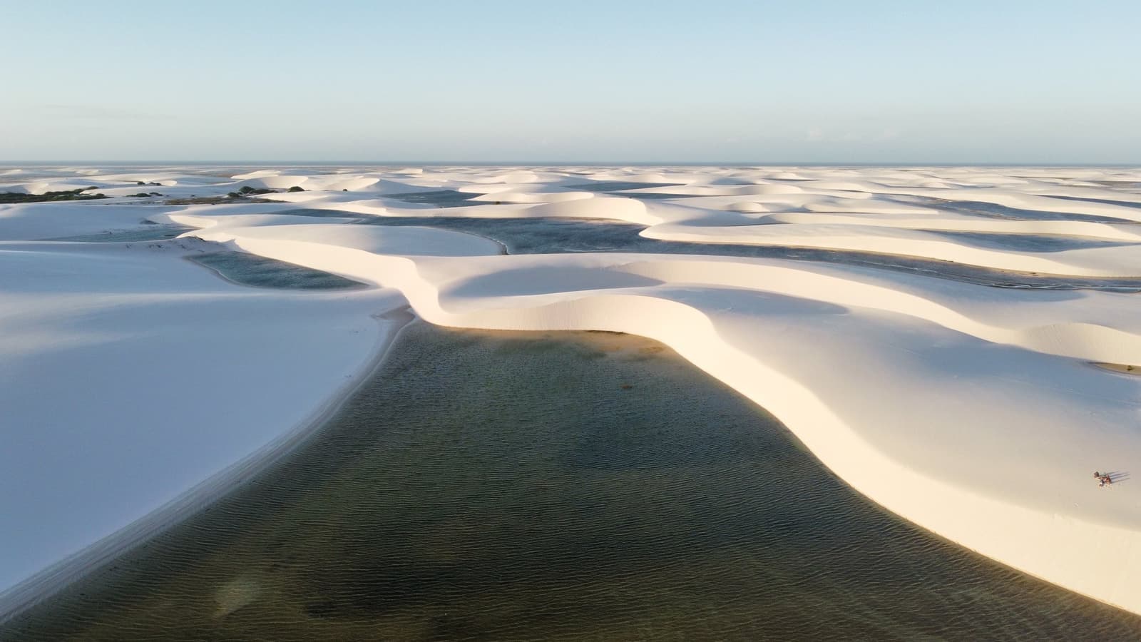 Sand dunes with their own lakes. Brazil keeps the best surprises. Aerial view of the Lençóis Maranhenses hike landscape, with sweeping white sand dunes curving around dark, clear freshwater lagoons stretching to the horizon under a pale blue sky