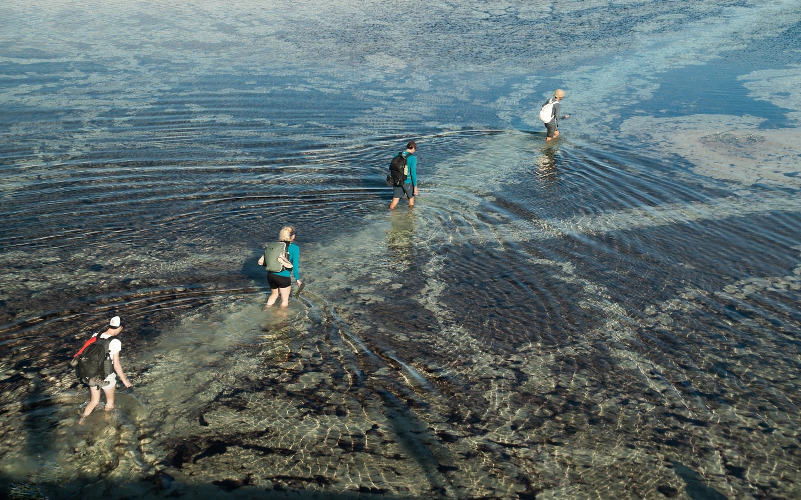 Wet feet are basically the entrance fee here. Four hikers wading through shallow, crystal-clear water on a Lençóis Maranhenses hike, their ripples spreading across a rocky riverbed as seen from above