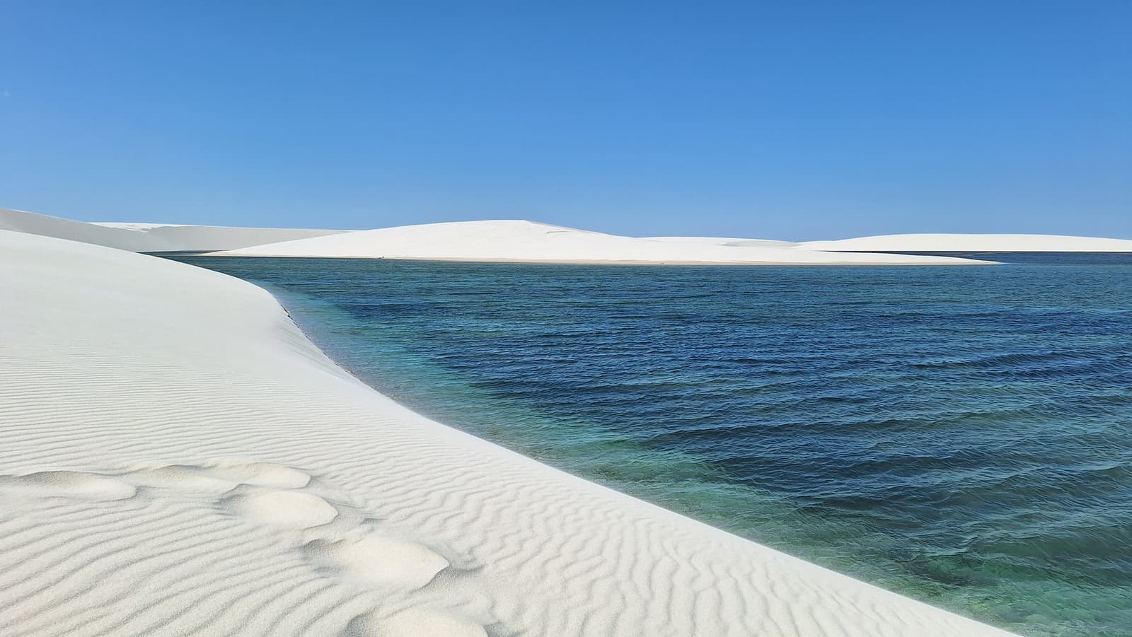 The lagoon is cold, the sand is hot, and the contrast never gets old. Rippled white sand dunes meeting a wide blue-green freshwater lagoon under a clear blue sky during the Lençóis Maranhenses hike, with more dunes rolling along the far shore
