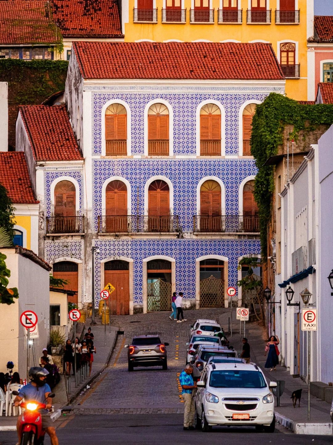 The azulejos do all the talking on this corner, the rest of the street just listens. A three-storey colonial building covered in blue-and-white Portuguese azulejo tiles on a cobblestone street in Santo Amaro, with colourful neighbouring facades, parked cars, and locals going about their day at golden hour