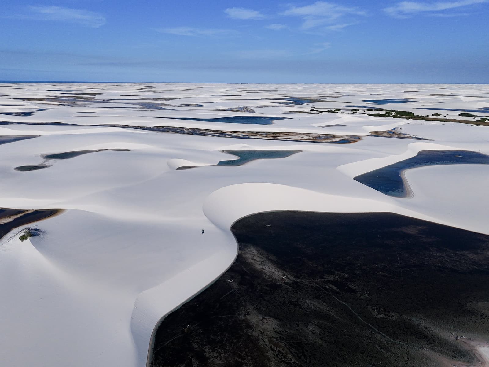 One person, a few million dunes. Scale has a way of humbling you here. Aerial view of the Lençóis Maranhenses Trek from Atins, showing vast white sand dunes stretching to the horizon, with dark and turquoise freshwater lagoons pooled between the dune ridges under a clear blue sky, and a lone figure visible on a dune crest in the middle distance