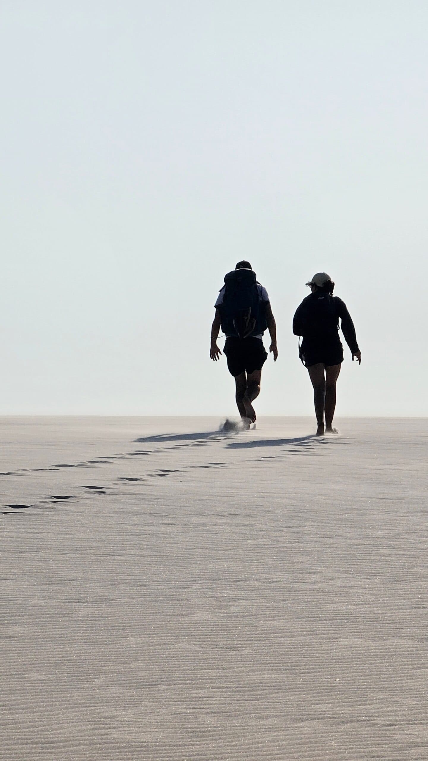 Just two people, an ocean of sand, and footprints that say you were here. Two trekkers walking away across a vast flat expanse of white sand dunes on the Lençóis Maranhenses trek from Atins, their footprints trailing behind them into a pale hazy sky