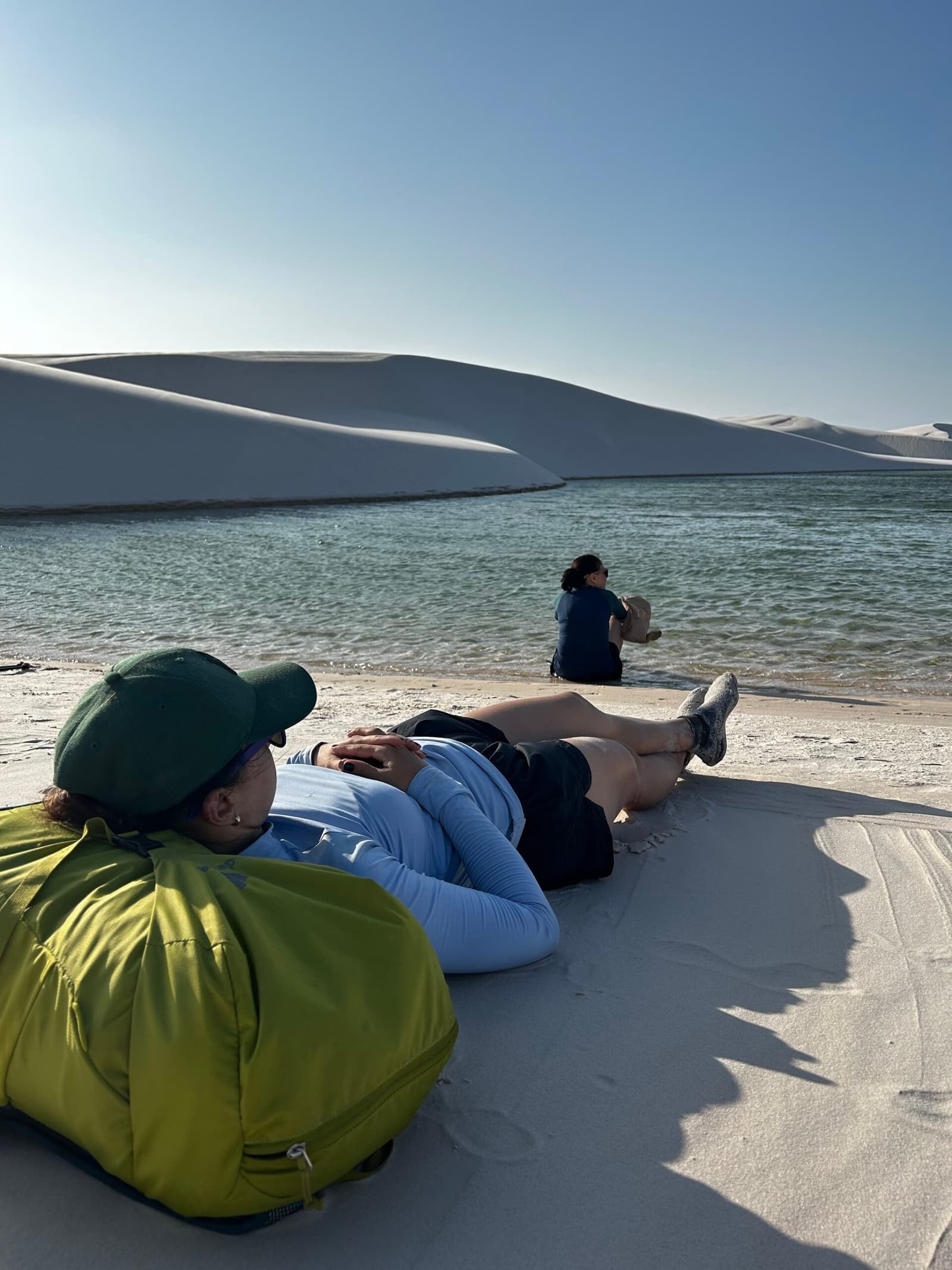 The dunes will wait. Sometimes the best move is just lying down. A traveler lying on white sand dunes beside a clear green lagoon during a Lençóis Maranhenses trekking trip, with a second person sitting at the water's edge and large white dunes rising behind