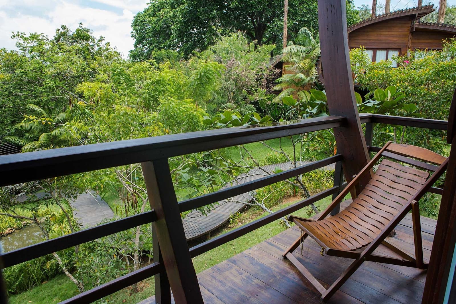 Your morning coffee spot, sorted. A wooden deck chair on a timber balcony at Mirante do Gavião overlooks a lush tropical garden with a boardwalk and neighbouring wooden bungalow