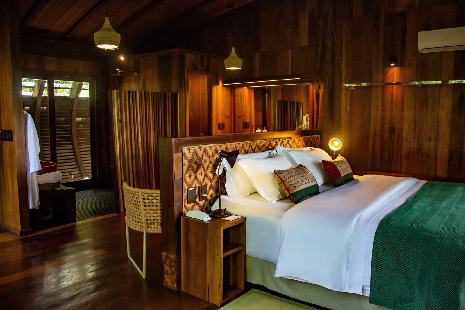 After a day on the river, this room earns its keep. A Mirante do Gavião amazon lodge suite featuring a king bed dressed in crisp white linen with hand-woven patterned cushions, a woven-rattan headboard, warm pendant lighting, and rich dark-timber walls opening toward a lush outdoor bathroom