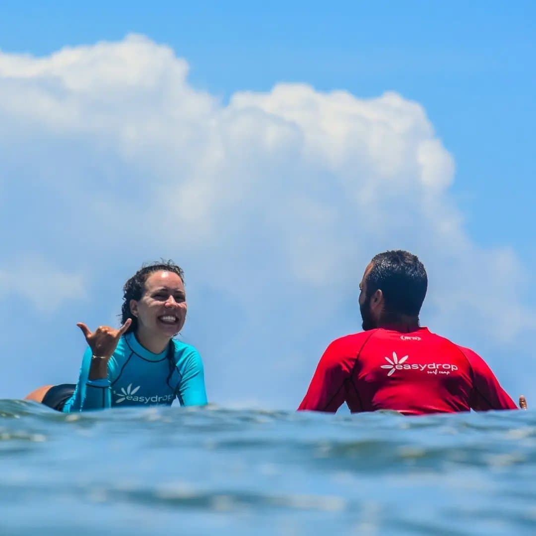That shaka says more than any score ever could. A smiling student in a teal EasyDrop rash guard flashes a shaka sign to her instructor during surf lessons in Itacaré, both sitting on their boards in the open ocean under a bright blue sky.
