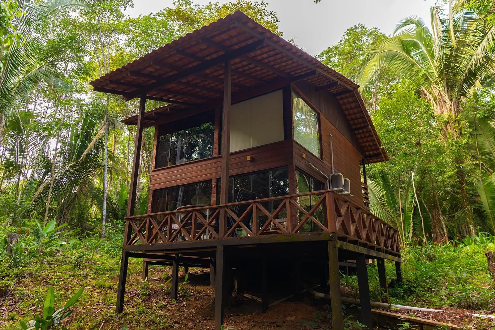 Raised above the forest floor, this bungalow keeps the jungle close without letting it in. A two-storey wooden Amazon boutique lodge cabin raised on stilts, with floor-to-ceiling glass windows and a wraparound deck, surrounded by dense tropical forest
