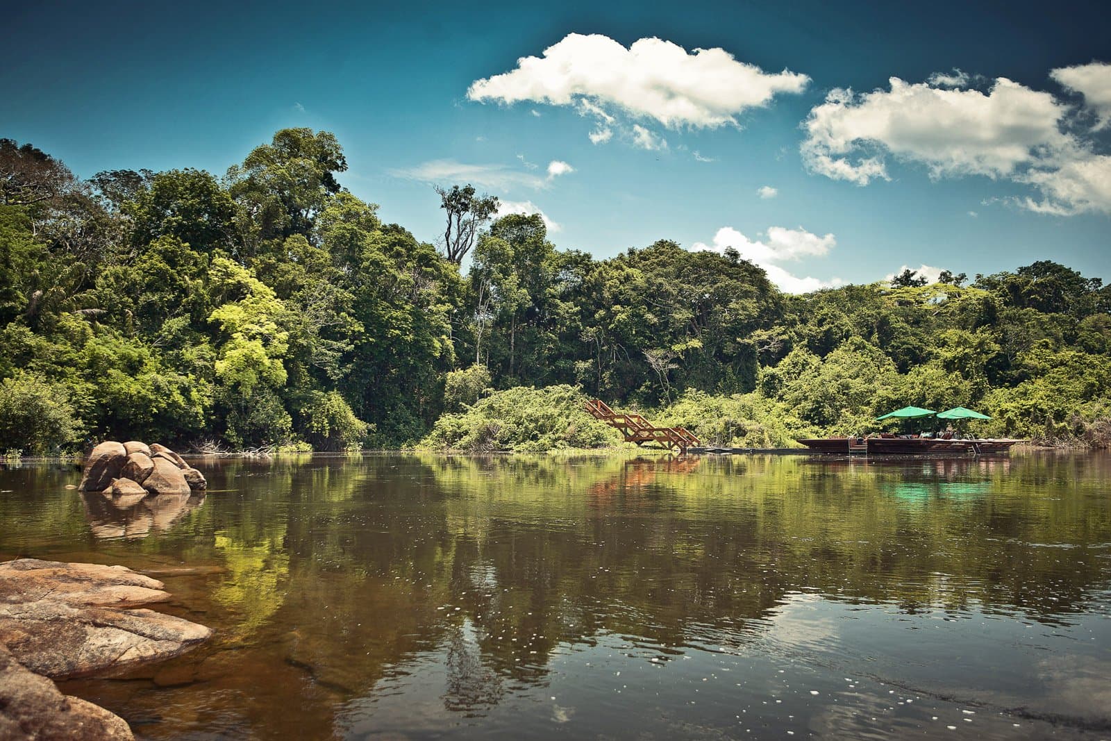 The river is your driveway. Everything else comes after. Photo: Samuel Melim (c) A calm dark-water river reflects the dense Amazon rainforest canopy, with granite boulders on the near bank, a wooden staircase landing, and two green umbrellas shading a dock on the far shore — the arrival point for birdwatching in the Amazon rainforest at Cristalino Jungle Lodge