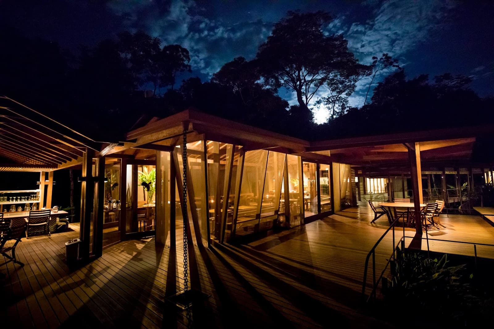 Dinner done, the forest dark, and the bar still open. Not bad. Cristalino Jungle Lodge restaurant and deck illuminated at night, warm amber light glowing through glass walls, forest canopy silhouetted against a moonlit sky — a comfortable base for birdwatching in the Amazon rainforest