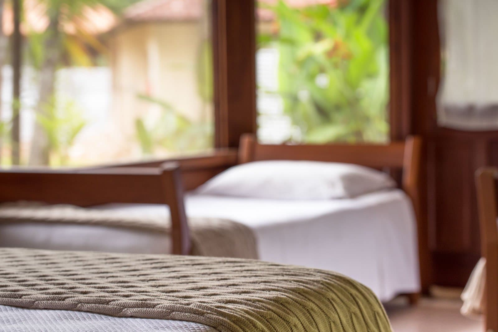 Crisp sheets, a knitted throw, and the forest just outside the glass. Photo: | SamuelMelim Photography Twin beds with wooden frames and a knitted green throw in a lodge room, large windows opening onto tropical garden foliage — comfortable accommodation for birdwatching in the Amazon rainforest
