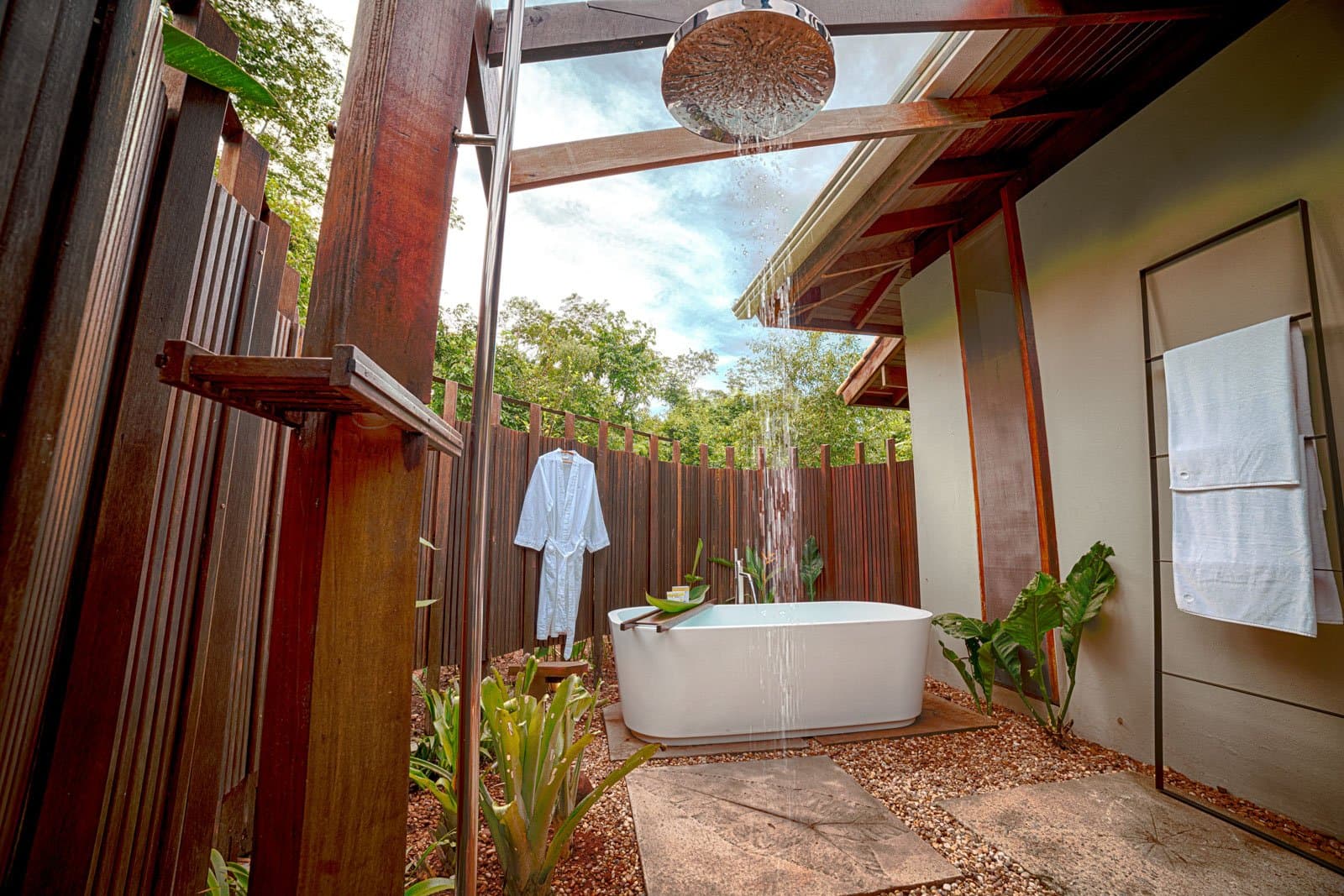 A rainfall shower open to the canopy, because some comforts just hit different in the forest. Photo: Samuel Melim Open-air bungalow bathroom at a birdwatching in the amazon rainforest lodge, featuring a freestanding white soaking tub beneath a rainfall showerhead, dark hardwood slatted walls, tropical plants, a hanging white robe, and a sky view through the open roof
