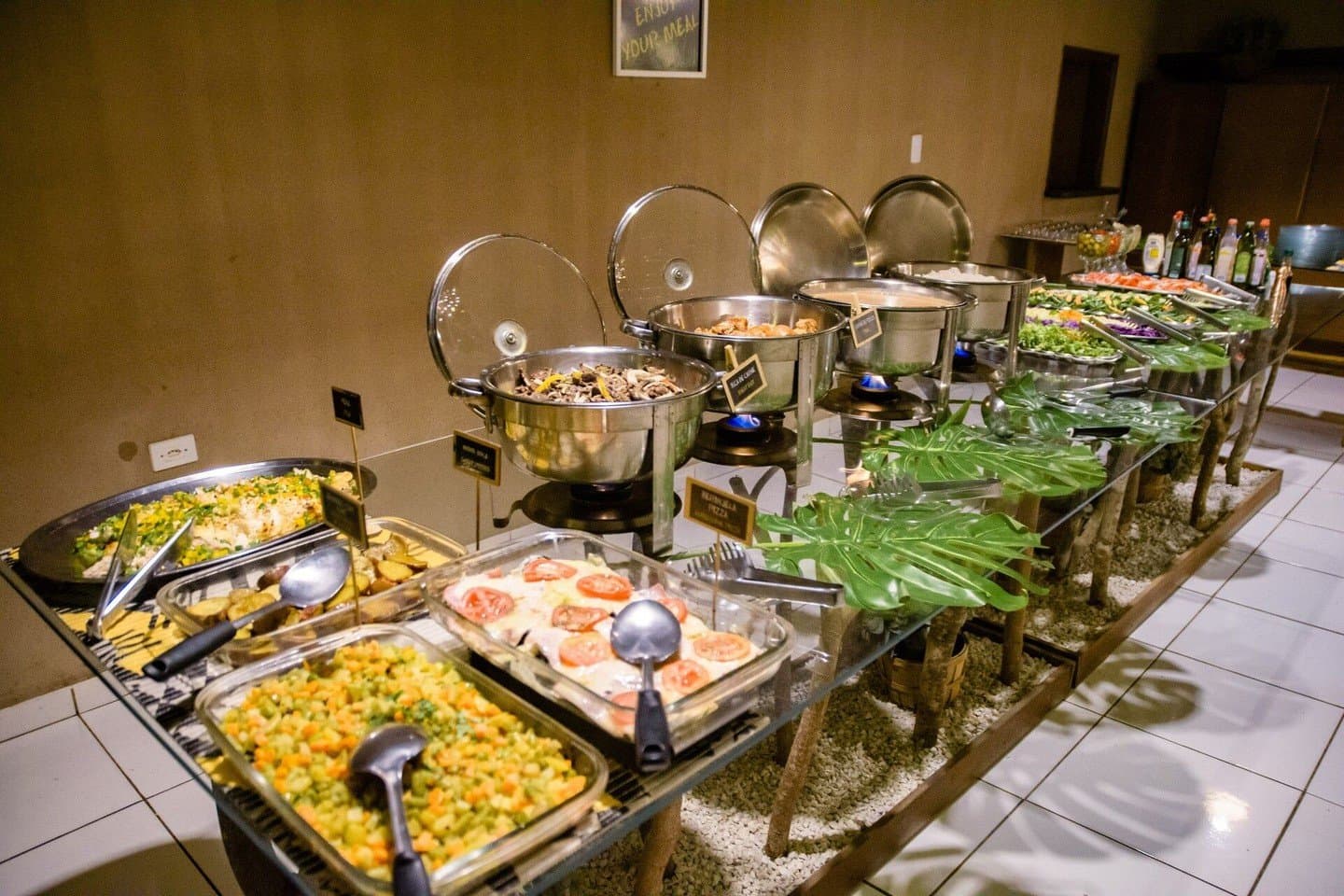 Turns out roughing it in the Pantanal still comes with a proper spread. A long buffet table at a Pantanal budget lodge spread with chafing dishes of hot food, a tomato-topped baked dish, diced vegetable salad, green salads, and condiments, decorated with tropical leaf accents