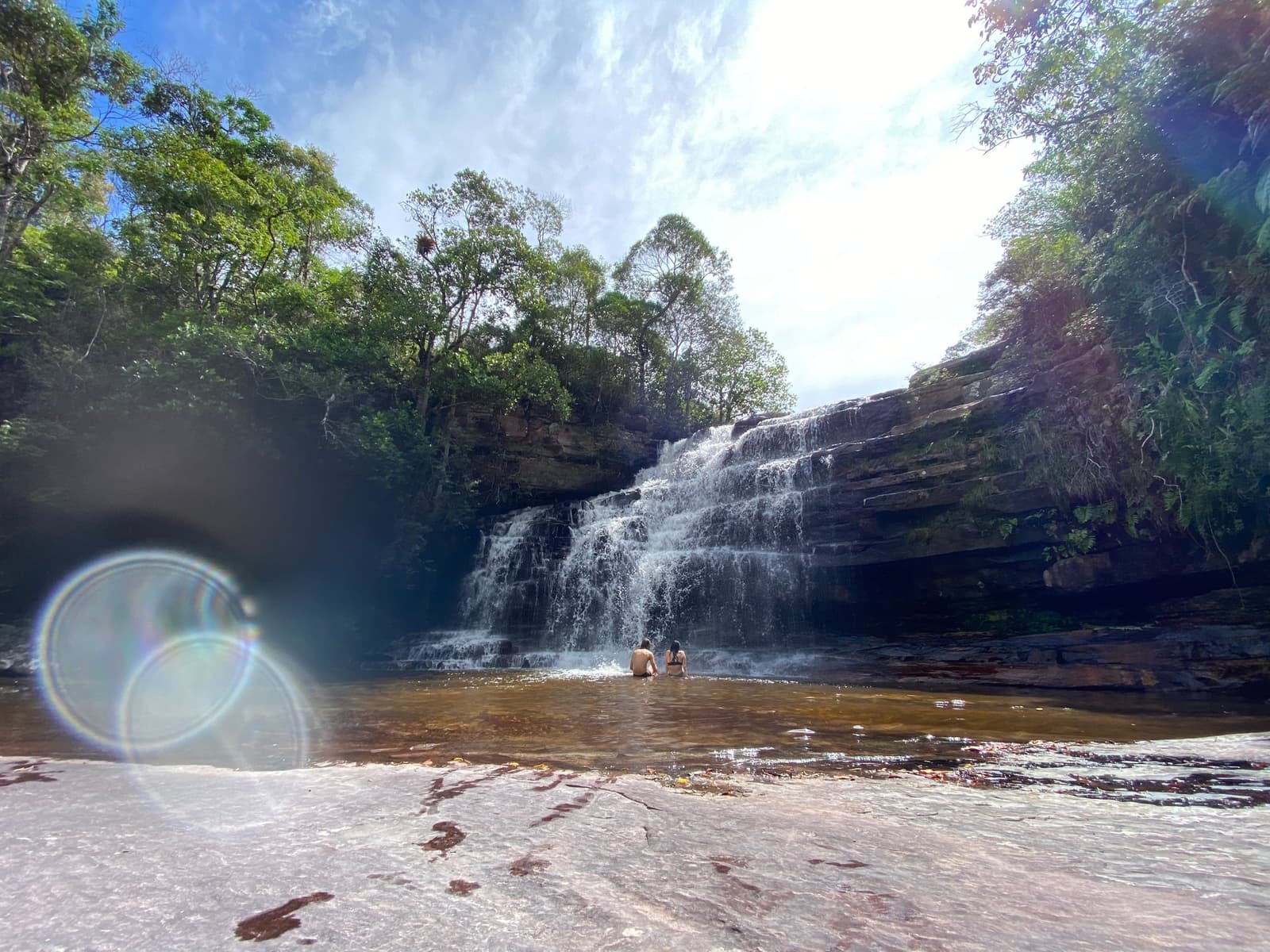 The pool is cold, the hike was earned, and nobody is leaving soon. Two swimmers wading in a clear natural pool at the base of a wide multi-tiered waterfall cascading over sandstone ledges, surrounded by green cerrado trees on a Chapada Diamantina trek