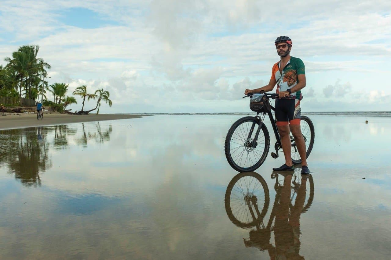 Wet sand, a perfect mirror, and forty kilometres still ahead of you. A cyclist in a green and orange jersey stands with his mountain bike on a flat, mirror-like wet beach, palm trees and cloudy sky reflected in the sand, with a second rider visible in the background — part of a cycling Bahia tour along the southern coast.