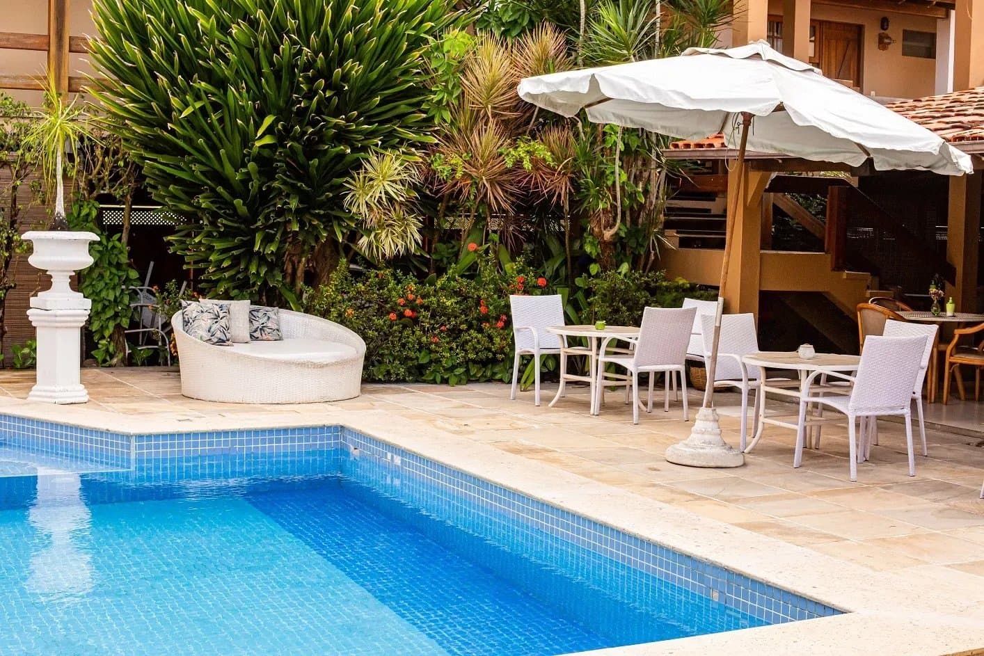 Cold water after warm roads. This pool earns every metre you pedalled. Sunlit pool terrace at a guesthouse on the Bahia cycling route, with white wicker lounge furniture, outdoor dining tables under a white umbrella, and lush tropical garden