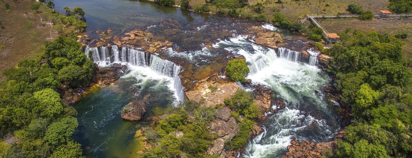 Twelve kilometres on foot, and then this shows up around the bend. Aerial view of a wide horseshoe-shaped waterfall on a rocky plateau surrounded by cerrado vegetation, with a small viewing platform visible on the right bank, seen during a Jalapão trekking route