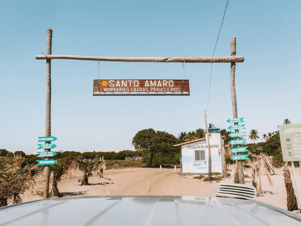 You know the adventure is real when the welcome sign lists lagoons, beaches, and rivers. Wooden welcome arch for Santo Amaro reading 'Morrarias Lagoas Praias e Rios', with turquoise directional signs and a sandy unpaved road ahead, part of the Lençóis Maranhenses full itinerary