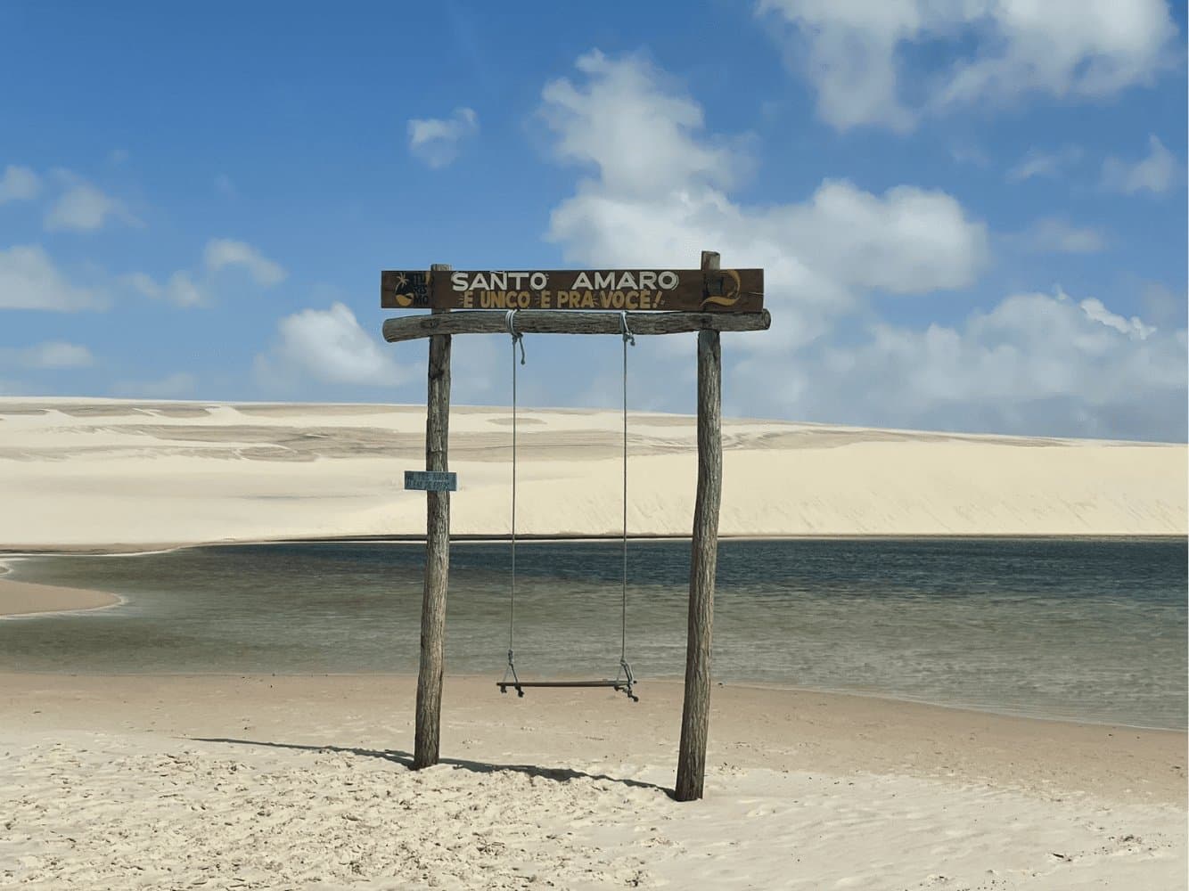 Santo Amaro knows it's special, and it's not shy about saying so. A rustic wooden swing and sign reading 'Santo Amaro é único e pra você' stand on white sand beside a calm lagoon, with sweeping white dunes stretching to the horizon on this Lençóis Maranhenses full itinerary stop