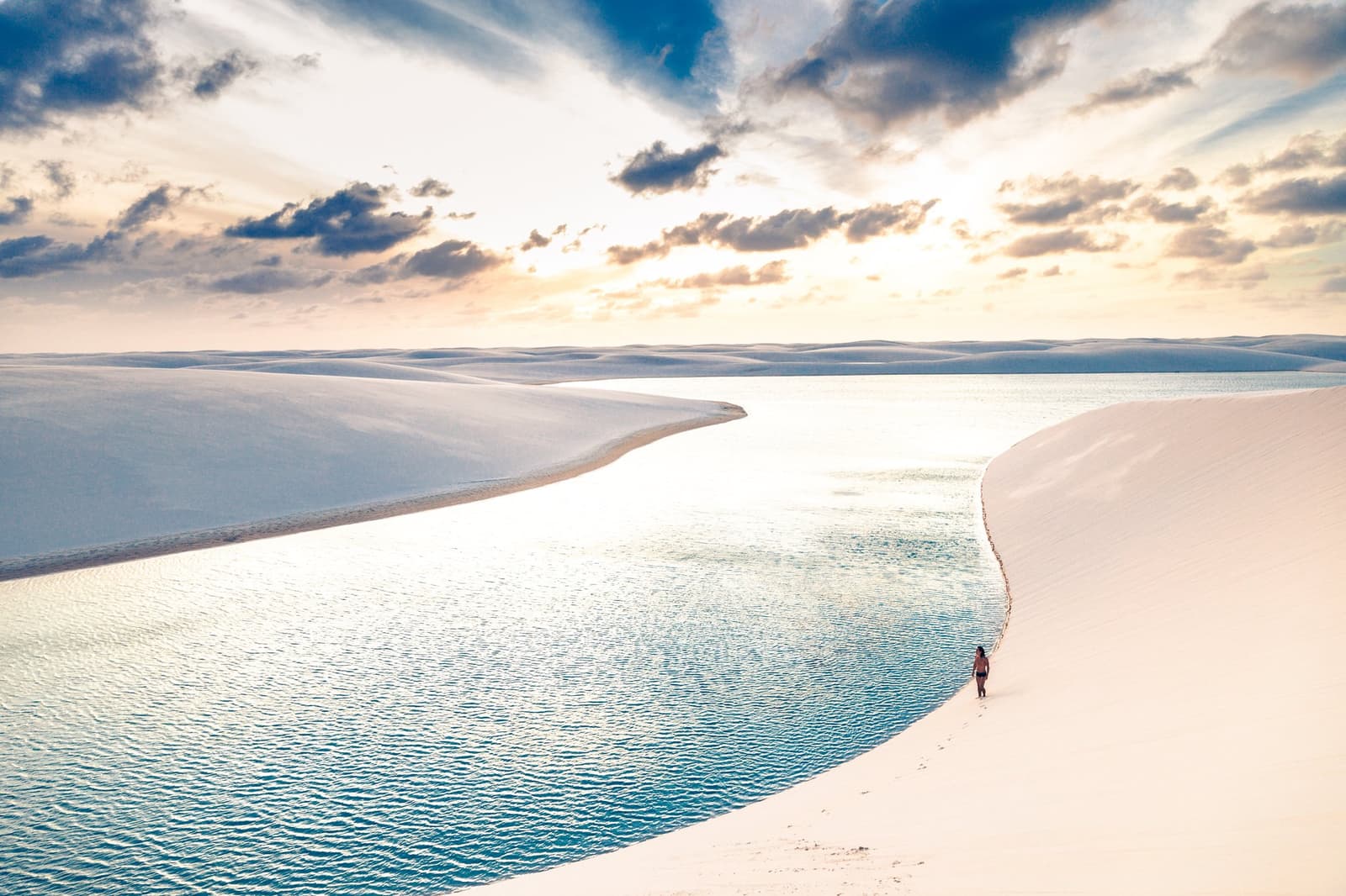 Just you, white sand, and a lagoon that appeared out of nowhere. A lone hiker walks along the curved edge of a glassy freshwater lagoon nestled between white sand dunes under a golden cloudy sky on the Lençóis Maranhenses hike