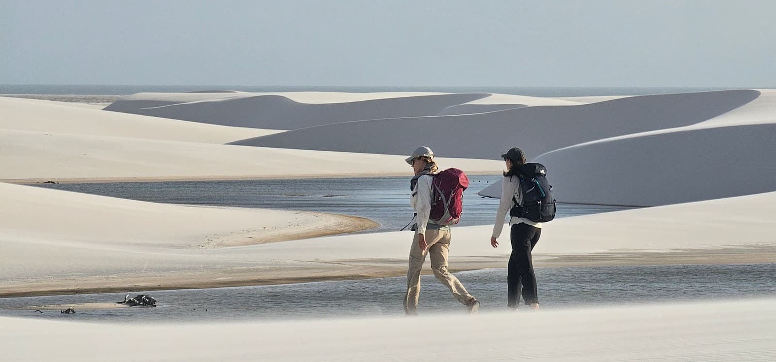 Good conversation makes the dunes feel half as long. Two hikers with backpacks walking across the white sand dunes of the Lençóis Maranhenses trek, with a shallow lagoon and sweeping dune ridges stretching to the horizon behind them