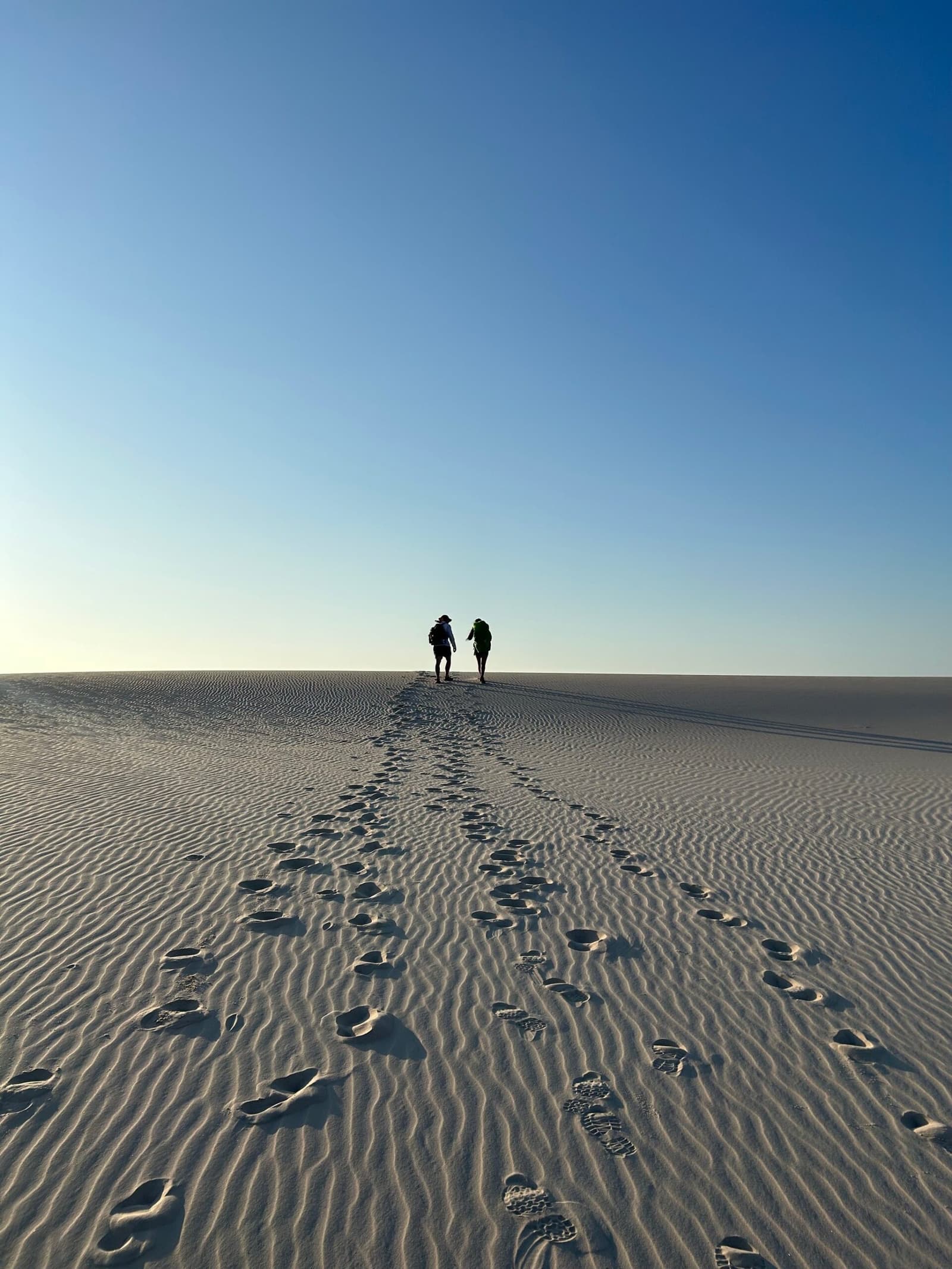 Two sets of footprints, one destination, zero wrong turns so far. Two hikers with backpacks silhouetted against a clear blue sky on a vast rippled white sand dune during a Lençóis Maranhenses trekking crossing, a trail of footprints stretching behind them