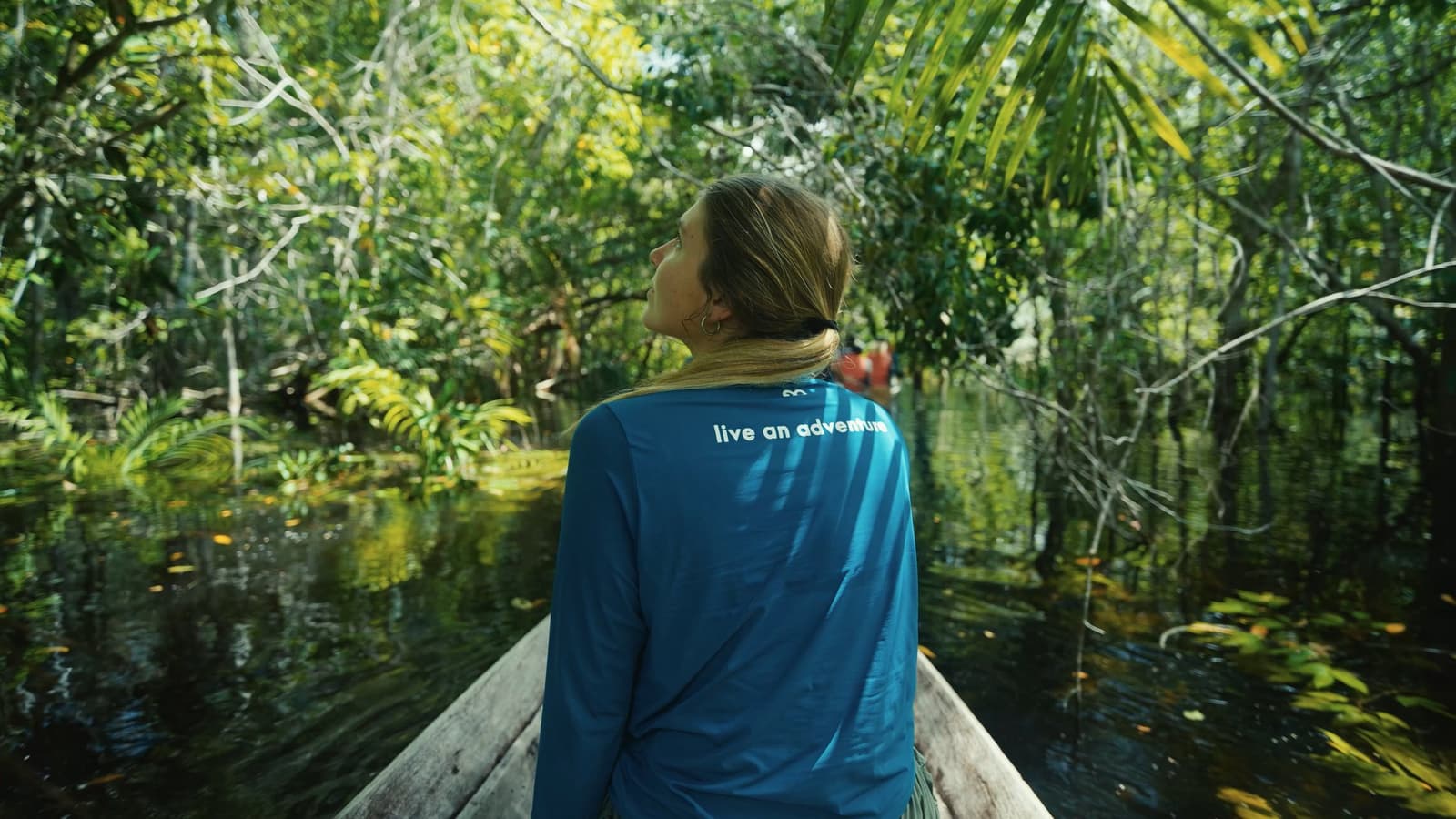 The forest doesn't open up for you, you slip into it, one paddle stroke at a time. A traveller seated at the bow of a wooden canoe gazes upward into a dense flooded igapó forest on a Mirante do Gavião Amazon Lodge excursion, dark tannin-stained water reflecting the lush green canopy overhead