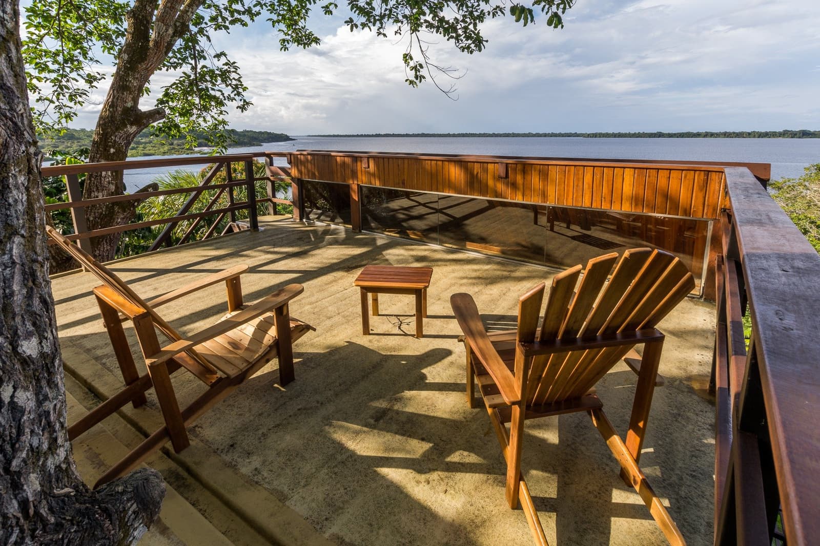 Your spot. The river decides the agenda. Two wooden Adirondack chairs and a small table on an open timber deck at Mirante do Gavião amazon lodge, overlooking a wide dark-water river bordered by dense rainforest canopy