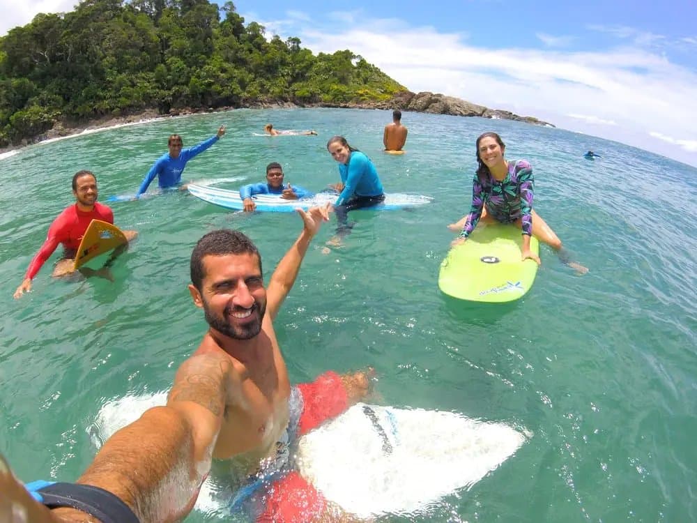Five boards, a lot of smiles, and nobody thinking about work right now. A smiling instructor takes a wide-angle water selfie with a group of students during surf lessons in Itacaré, all holding colorful surfboards in clear turquoise water with a forested headland behind them.