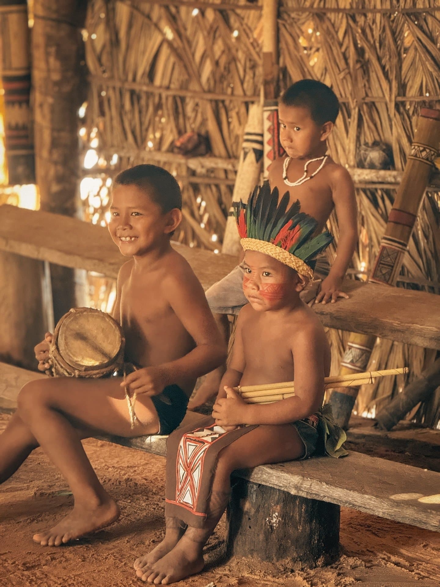 The music starts young, and these kids clearly know it. Three indigenous children in traditional face paint, feather headdress and woven accessories sit inside a thatched communal hut during a village visit on an Amazon jungle cruise Brazil itinerary
