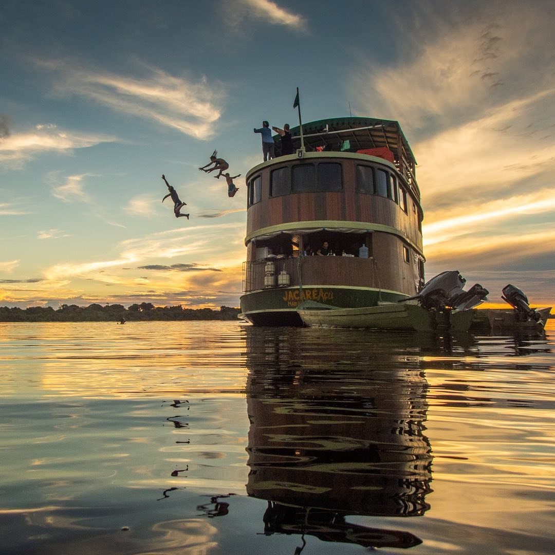 Jumping in is always the right call at the end of a good day. Guests leaping from the upper deck of the Jacareagil riverboat into a glassy river at sunset on an Amazon river cruise in Brazil, with golden light reflected across the calm black water