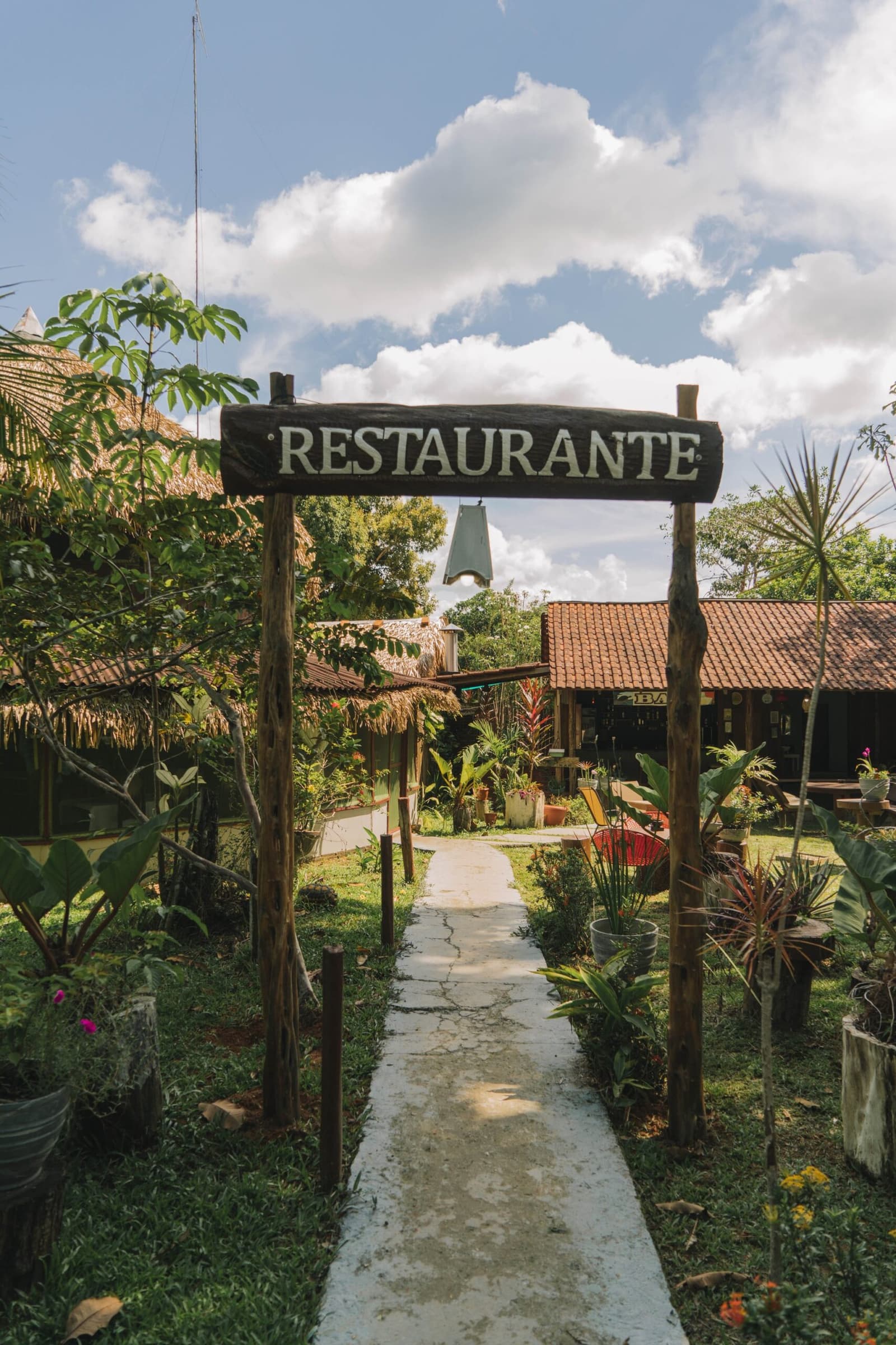 Good meals taste better when the walk there looks like this. A rustic wooden 'Restaurante' sign arching over a stone pathway leading through a lush tropical garden to the open-air dining area at the Amazon Turtle Lodge