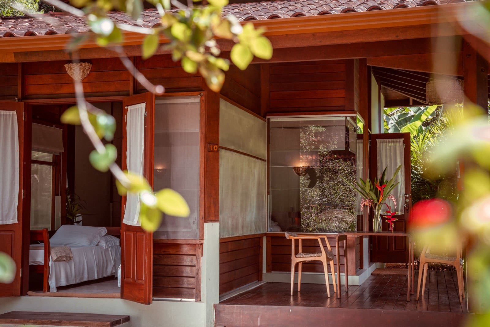 Room 10 keeps it honest: wood, white linen, and the jungle right outside. Photo: Samuel Melim Wooden bungalow at Cristalino Jungle Lodge with open shuttered doors revealing a white-sheeted bed inside, a covered veranda with timber chairs, and tropical foliage framing the facade — a comfortable base for birdwatching in the Amazon rainforest