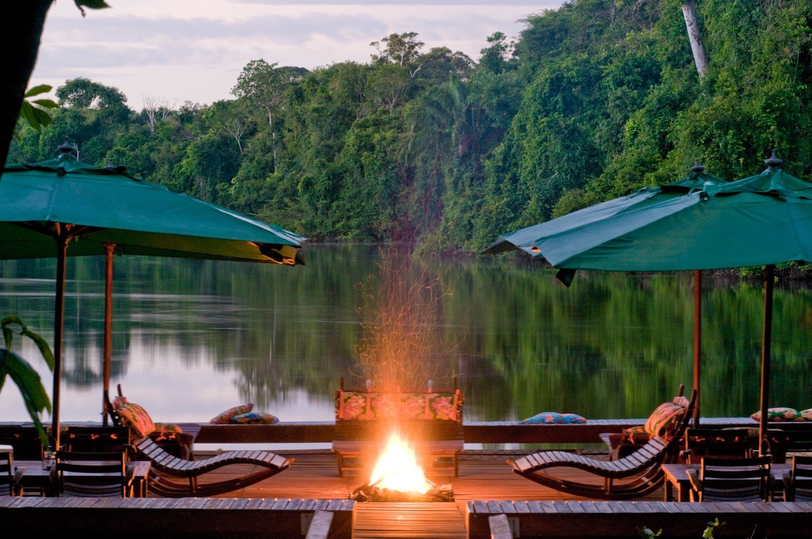 Firelight, still water, and the forest closing in. Not a bad way to end a long day on the trails. Photo: Luis Gomes A campfire burning on a wooden deck at dusk, surrounded by cushioned lounge chairs and green umbrellas, overlooking a calm river with dense rainforest reflected on the water — the ideal setting for birdwatching in the Amazon rainforest by day, and stargazing by night.