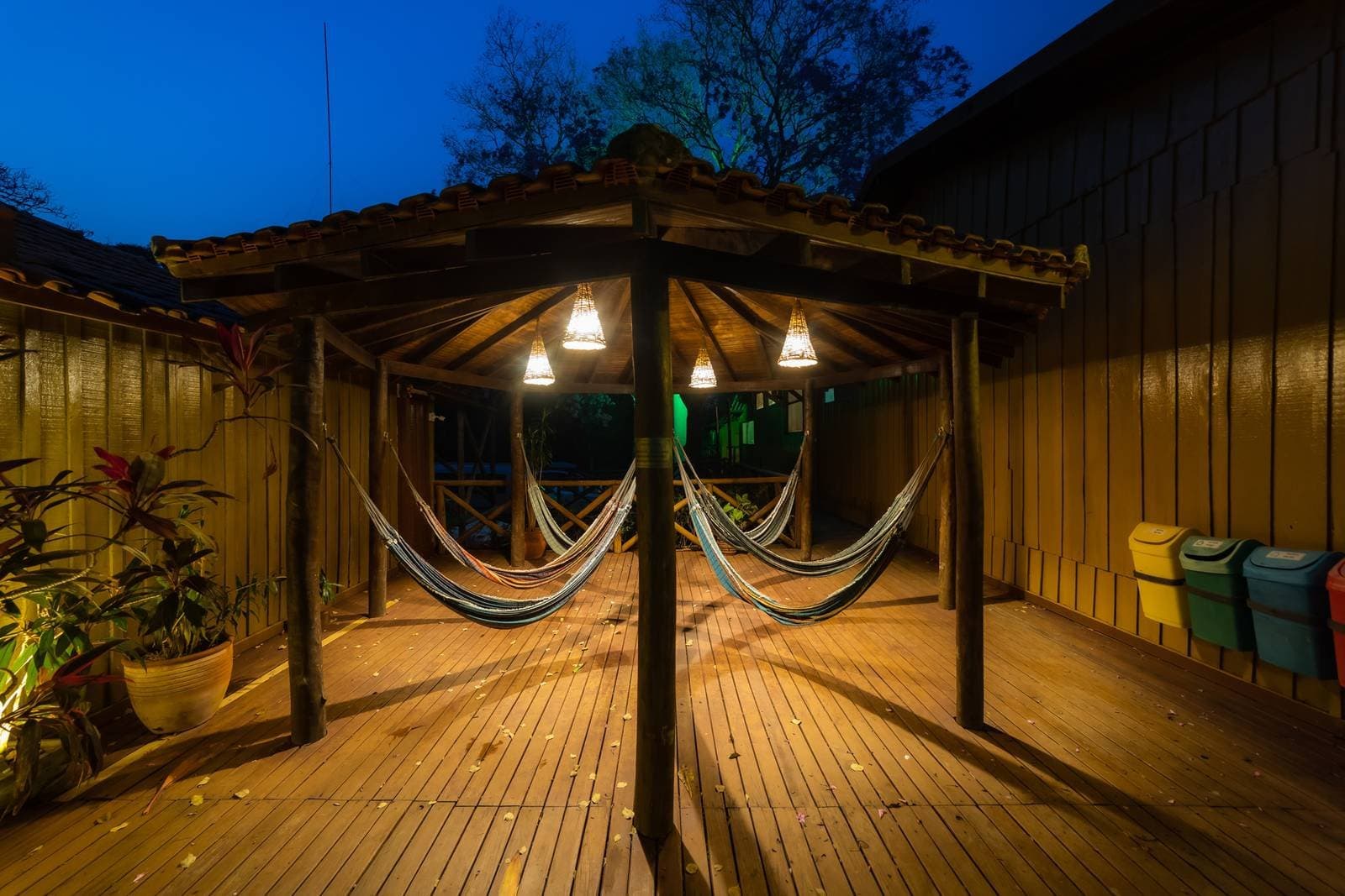 The kind of spot that makes you cancel tomorrow's early alarm. Covered wooden deck at dusk on a budget Pantanal tour, with multiple colorful hammocks hanging beneath warm pendant lights and a deep blue evening sky above the treeline