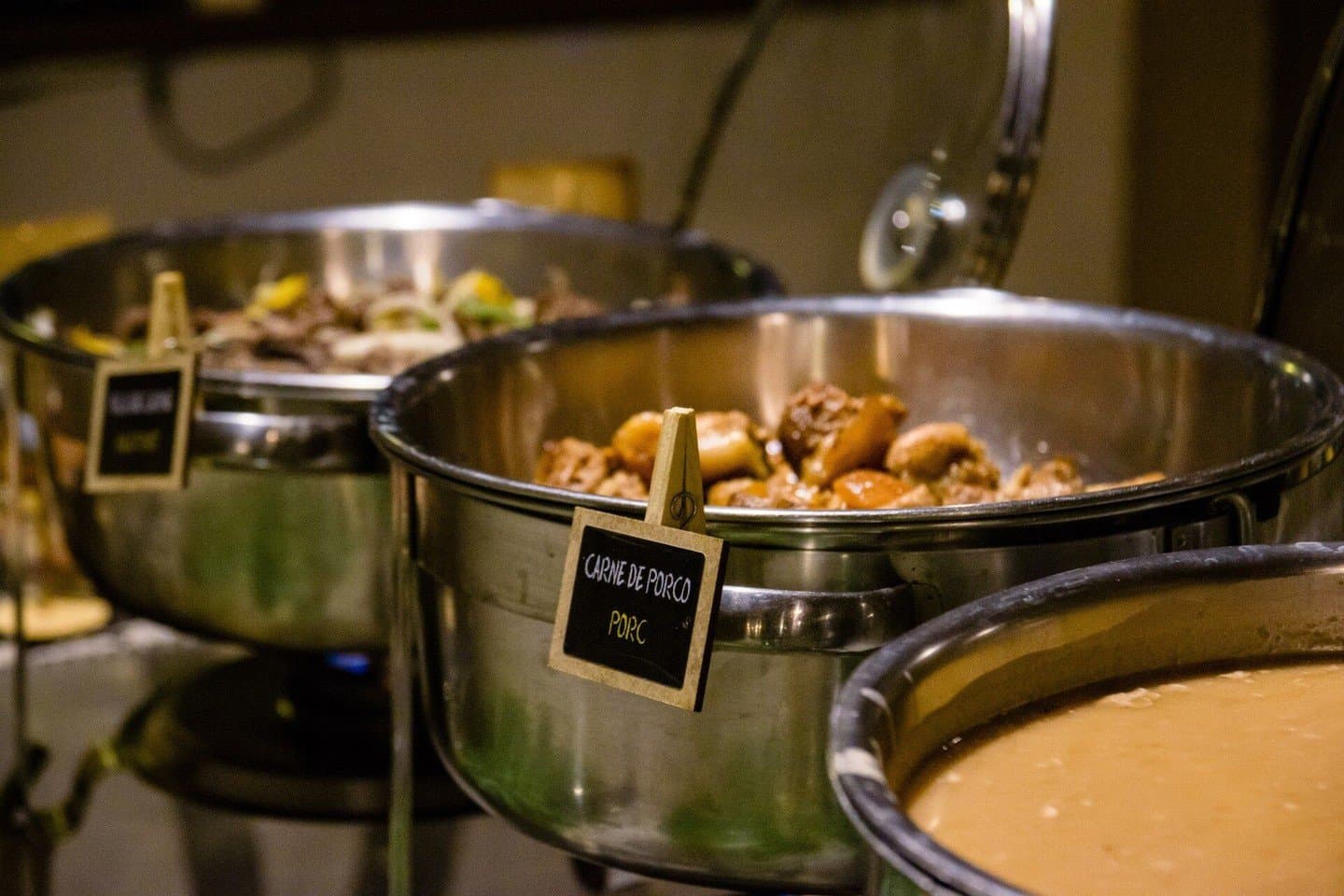 Honest, hearty food after a long day on the water. Stainless steel chafing dishes at a lodge buffet, with the foremost pot labeled 'Carne de Porco' holding braised pork pieces, part of the full-board meals included in a budget Pantanal tour