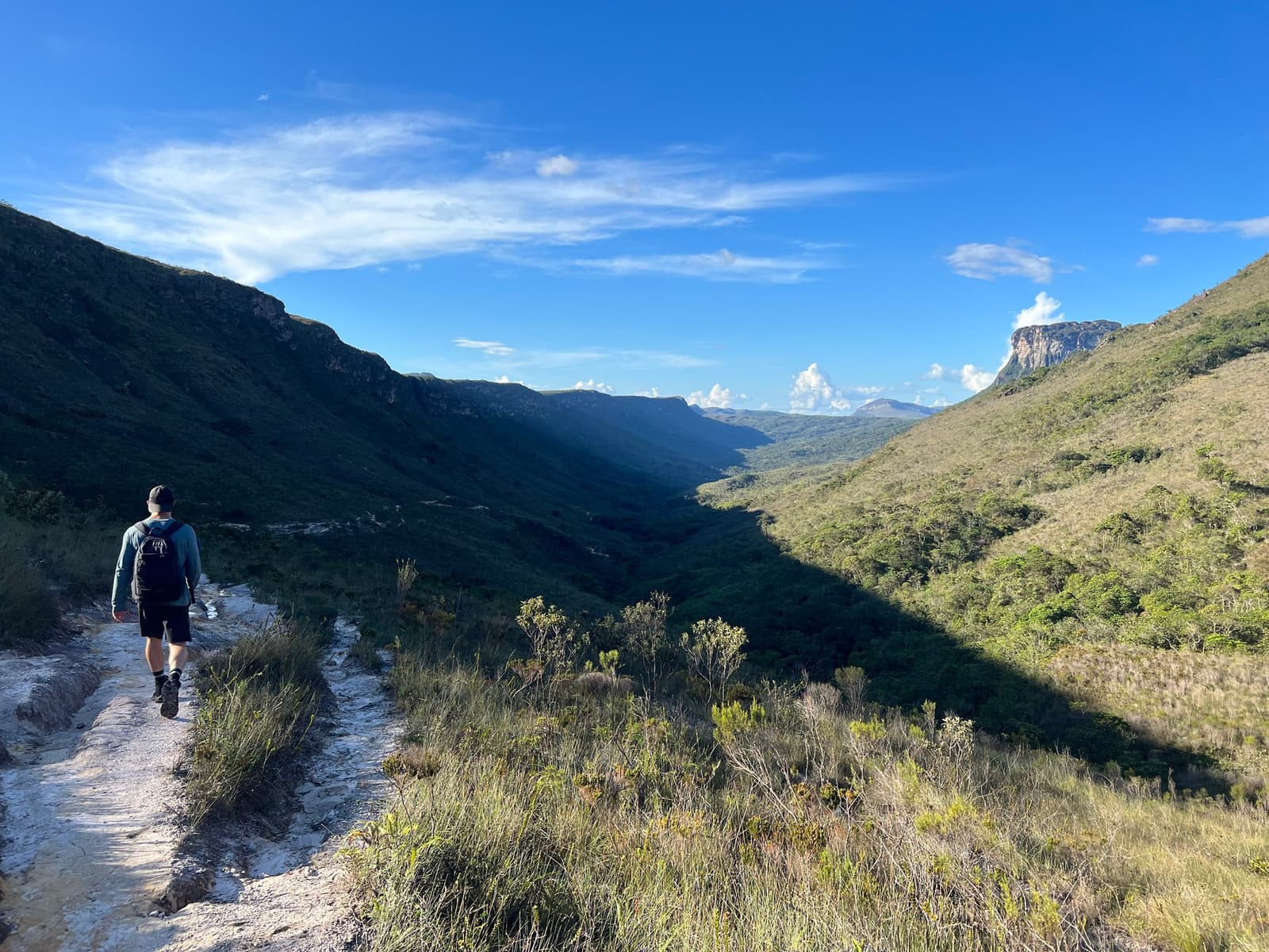The trail keeps going, and somehow, so do you. A hiker with a backpack walks a rocky trail on the Chapada Diamantina trek, with a wide green valley flanked by sandstone escarpments stretching into the distance under a blue sky