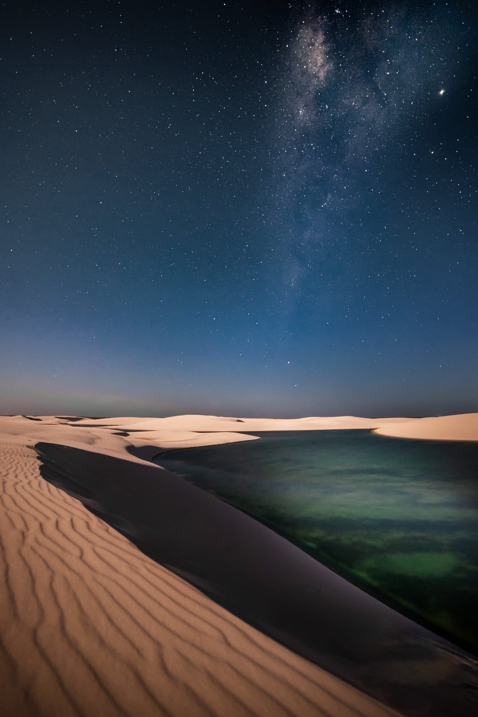 No light pollution, no noise, just the galaxy doing its thing above the dunes. The Milky Way arching over white sand dunes and a still turquoise lagoon at night in Lençóis Maranhenses Full Itinerary stargazing experience