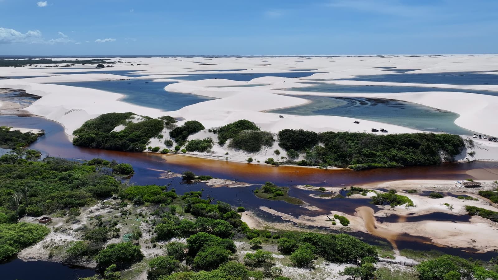 Two colours of water, one landscape, the dunes never quite follow the rules. Aerial view of a tannin-dark river winding through green vegetation before meeting the white dunes and turquoise lagoons of the Lençóis Maranhenses full itinerary landscape