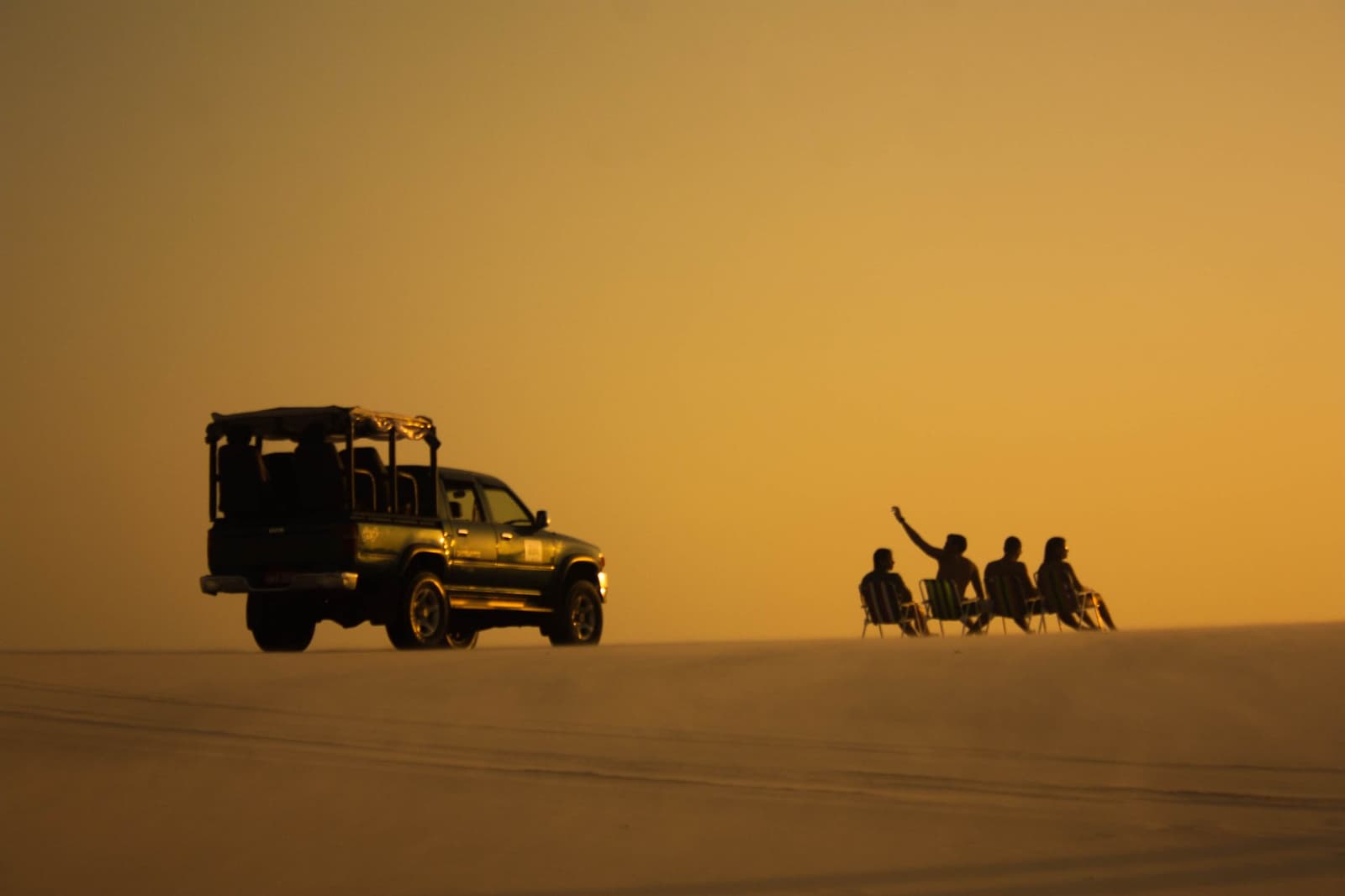 Chairs on the dunes, nowhere to be, and that light doing all the work. Four travelers seated in folding chairs on a vast open dune at golden-hour sunset, with a 4x4 safari vehicle silhouetted beside them during a Lençóis Maranhenses full itinerary excursion