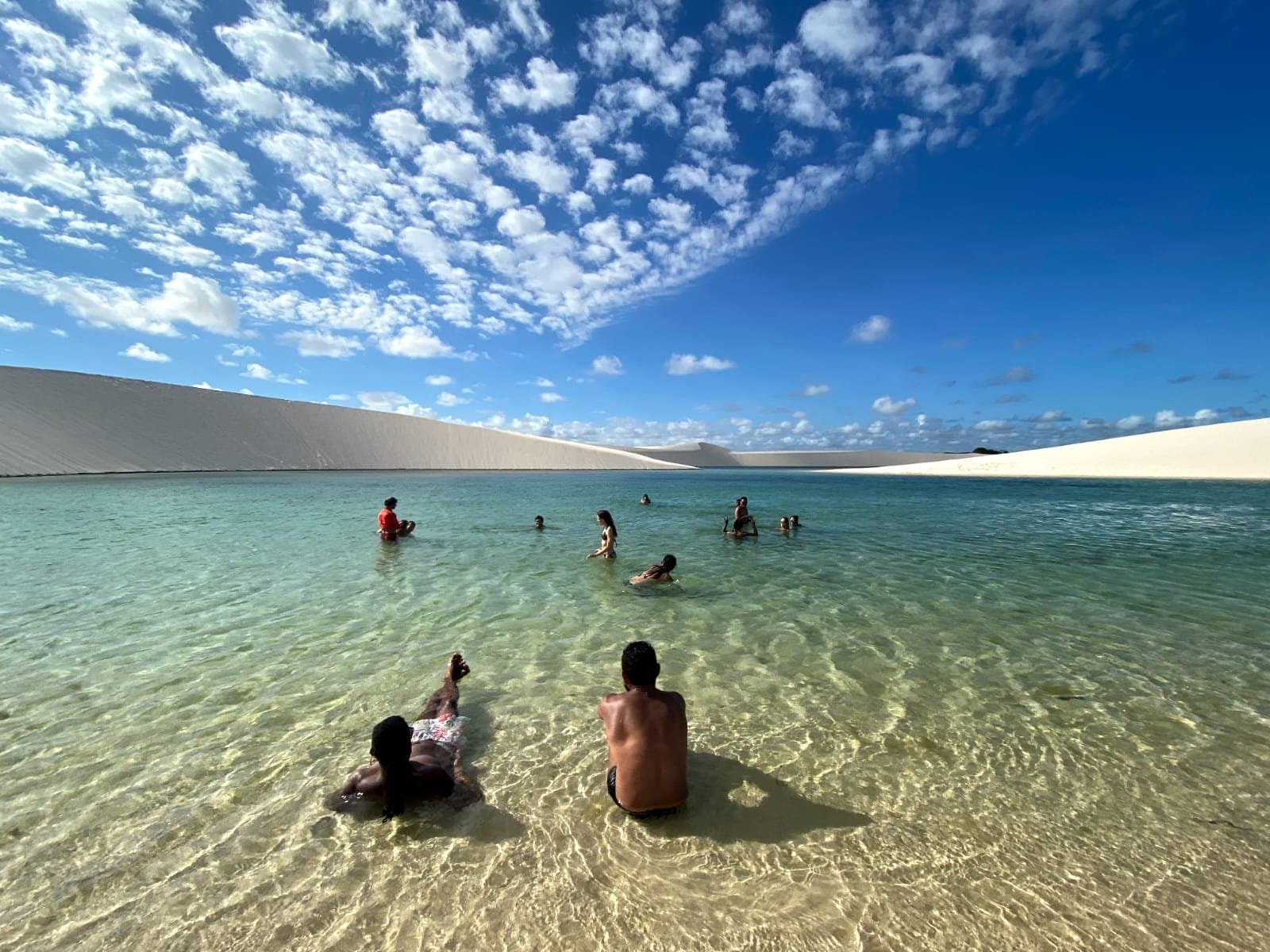 Cool, clear, and nowhere near a swimming pool. This is the real thing. Swimmers wading and floating in a clear turquoise freshwater lagoon during a Lençóis Maranhenses hike, with smooth white sand dunes curving along the far shore under a blue sky scattered with small clouds