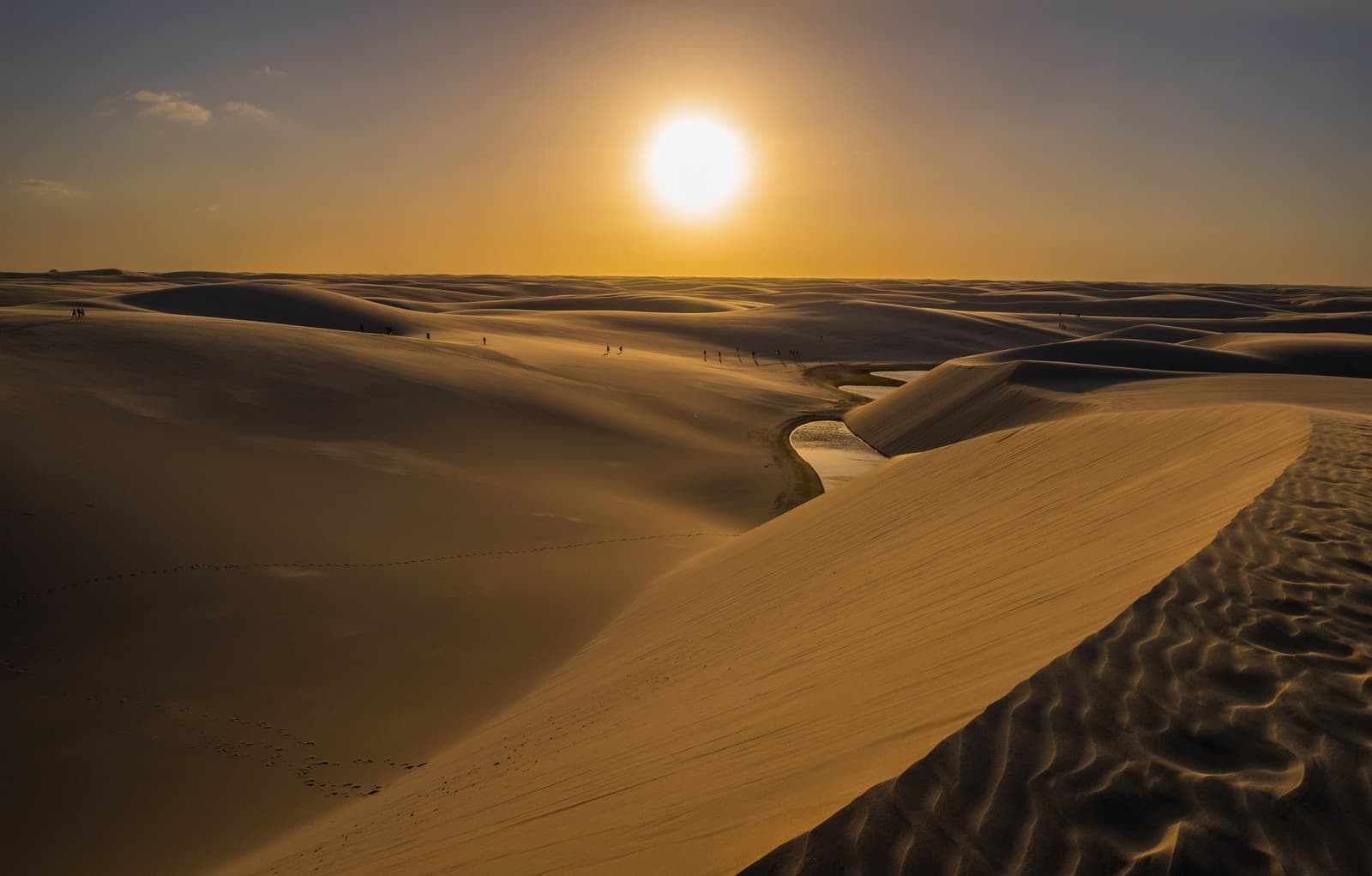 A desert that fills with water. The Lençóis Maranhenses plays by its own rules. Vast golden sand dunes of Lençóis Maranhenses stretch to the horizon at sunset, with a small freshwater lagoon curving between the dunes and tiny silhouettes of hikers visible in the middle distance during a Lençóis Maranhenses hike
