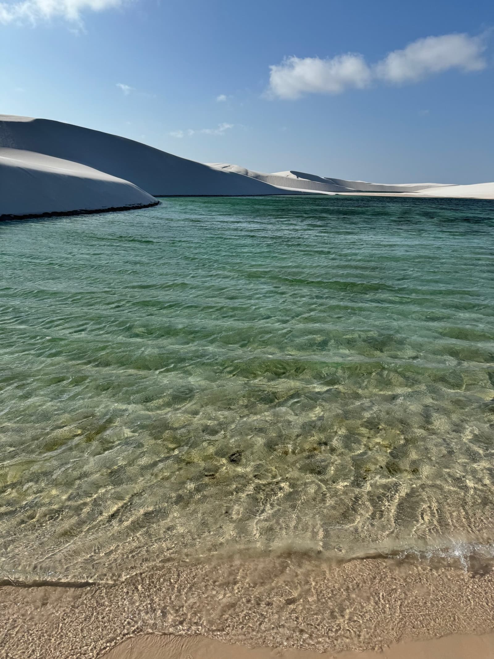 Cool, clean water and not a footprint ahead, this is the good part. Crystal-clear green water of a freshwater lagoon on the Lençóis Maranhenses trek, with towering white sand dunes rising along the far shore under a blue sky