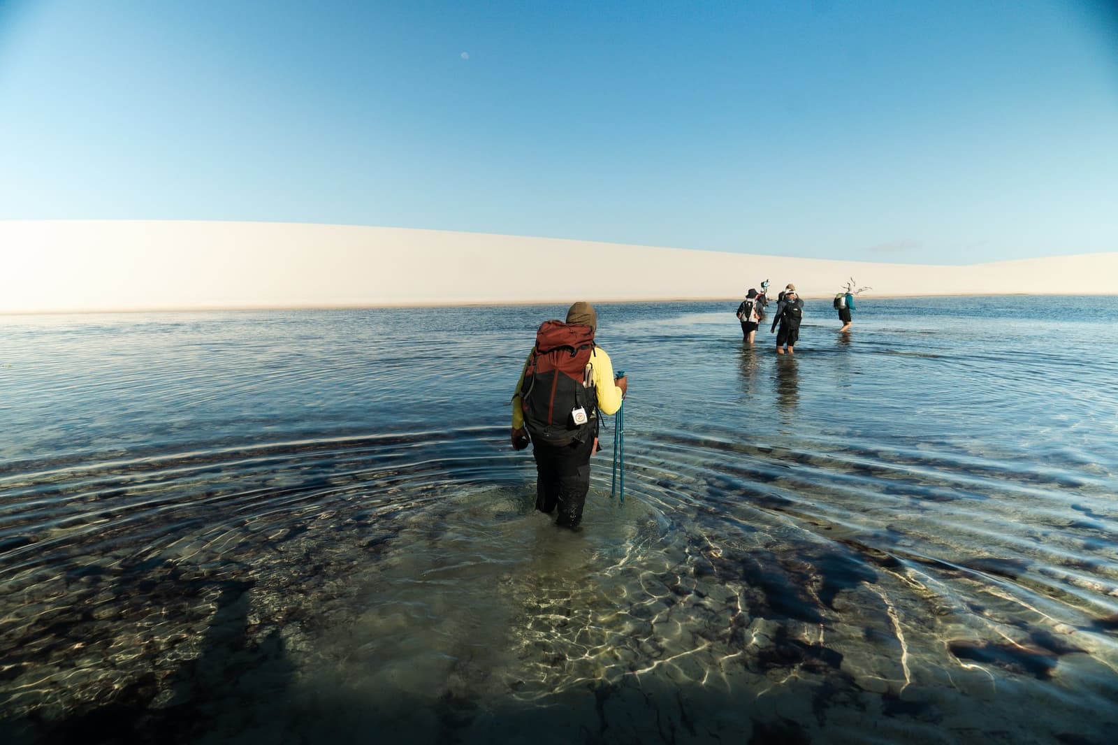 Boots off, packs on, some crossings are just part of the deal. A group of backpackers wading knee-deep through a clear, shallow lagoon on the Lençóis Maranhenses trek, with a long white sand dune ridge stretching across the horizon under a blue sky.