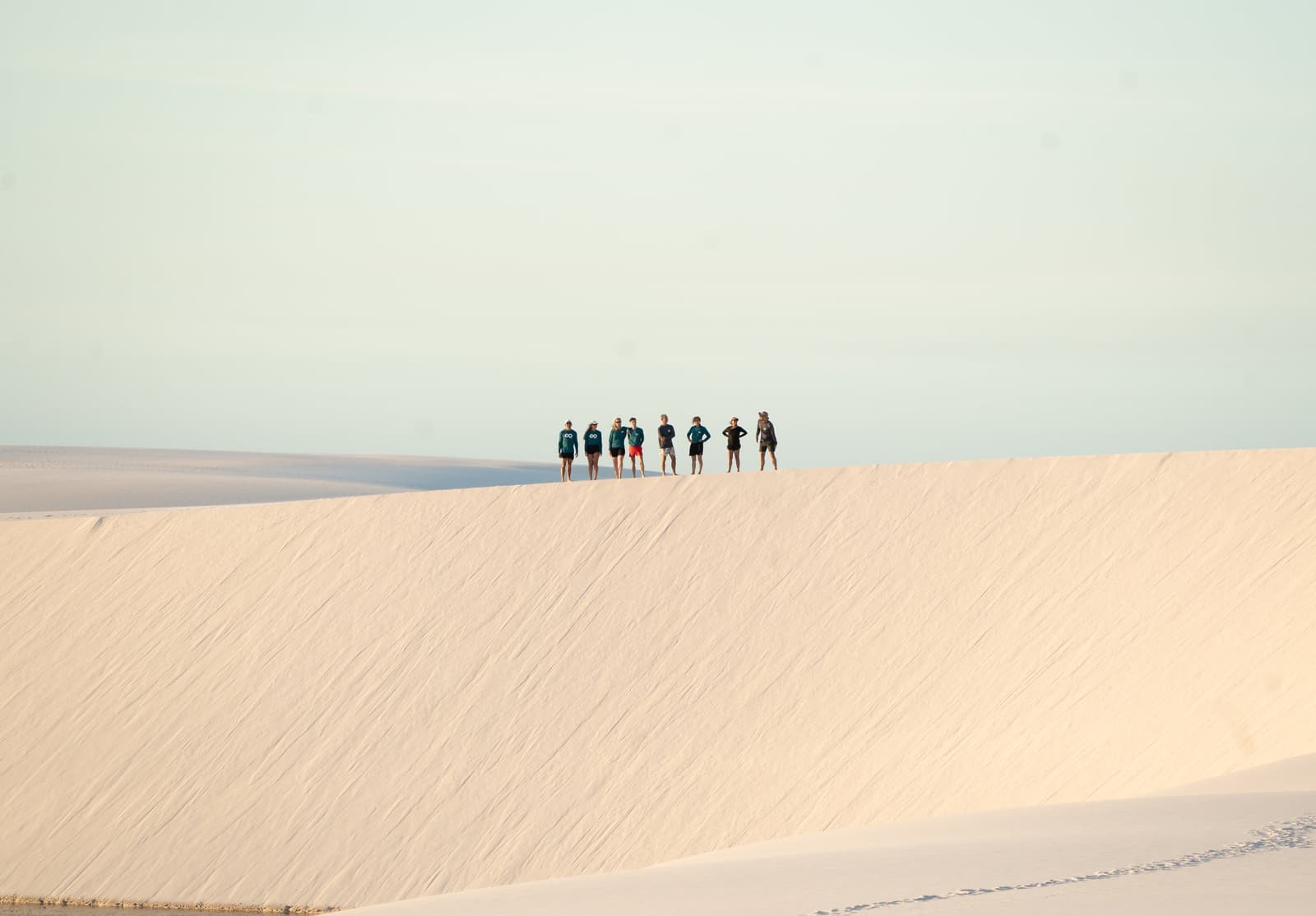 Seven people, one ridgeline, and an awful lot of sand between here and the horizon. A group of seven trekkers standing along the ridge of a vast white sand dune during a Lençóis Maranhenses trekking expedition, with a pale sky and endless dune landscape stretching behind them