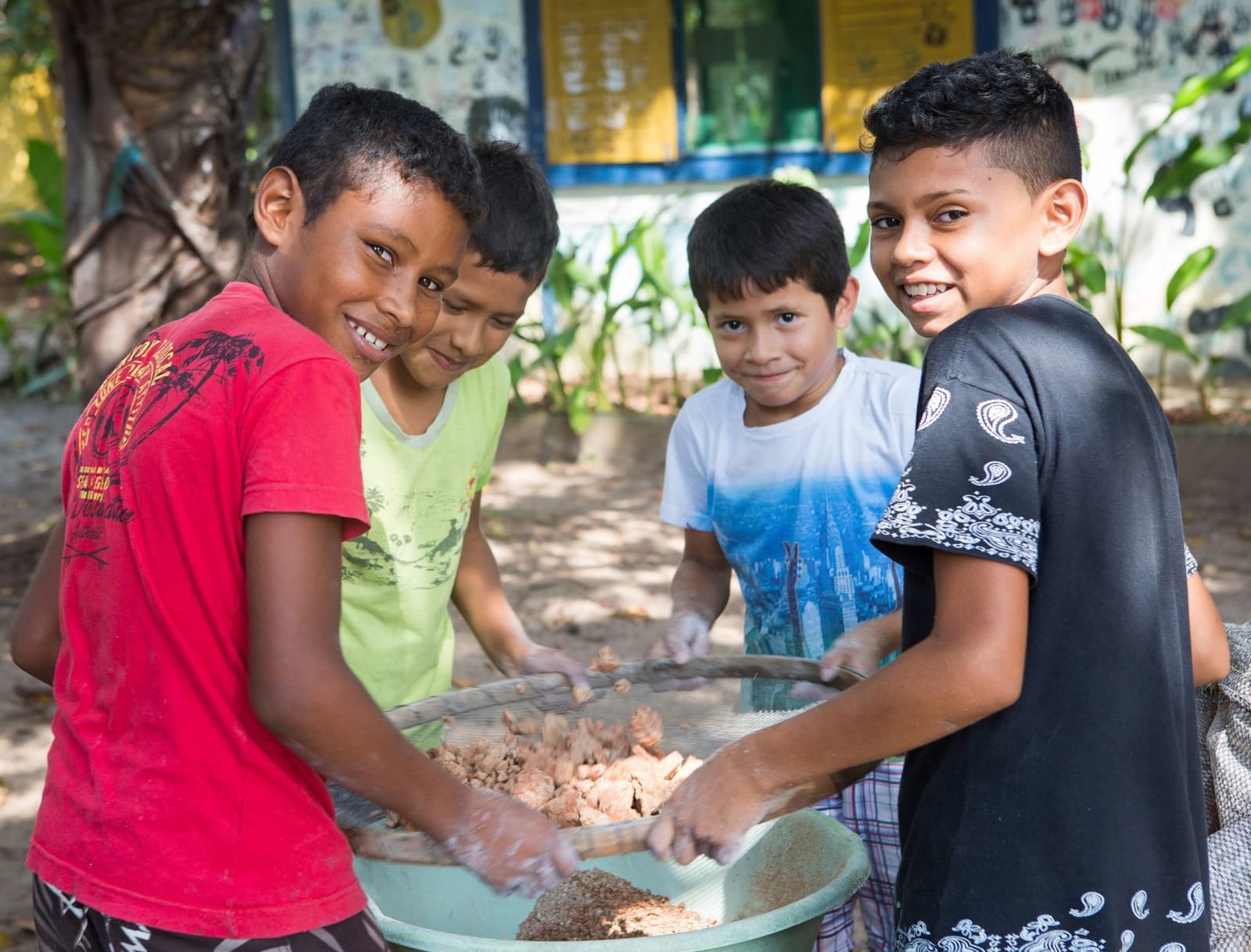 Getting their hands dirty is half the fun. Four smiling boys sieve and sort harvested food together at a riverside community near Mirante do Gavião, hands busy over a wide basin outdoors