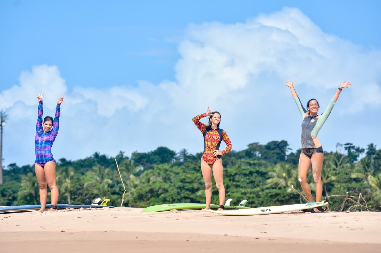 Three boards, three smiles, and the kind of energy that's contagious before you even hit the water. Photo: Picasa Three women in colorful rash guards standing on a sandy beach with surfboards at their feet, arms raised in celebration, at a surf school in Itacaré with tropical forest in the background