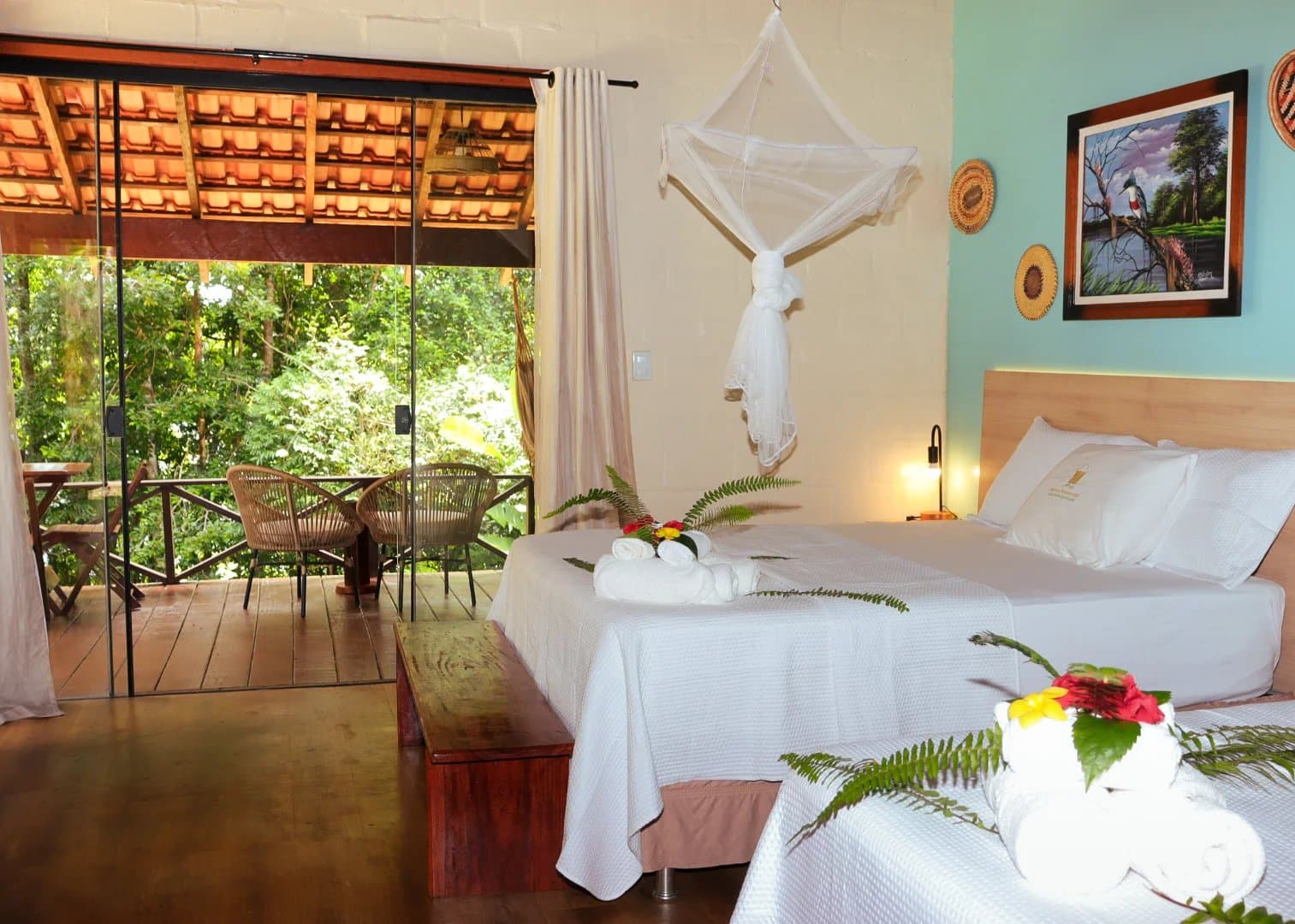 Crisp linens, a mosquito net, and the Amazon just a sliding door away. Interior of an Amazon boutique lodge room with two white beds dressed with folded towels and tropical flowers, a hanging mosquito net, warm bedside lighting, local artwork on the wall, and sliding glass doors opening onto a wooden balcony with rattan chairs and dense green forest beyond