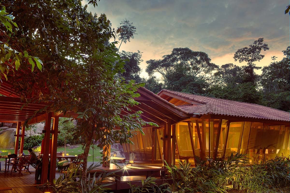 Candles on, forest close, and the river just beyond. This is where the day winds down. Photo: Samuel Melim Cristalino Jungle Lodge lit from within at dusk, its warm timber and glass pavilion surrounded by dense Amazon rainforest canopy and tropical garden, lanterns glowing on the wooden deck — a peaceful base for birdwatching in the Amazon rainforest