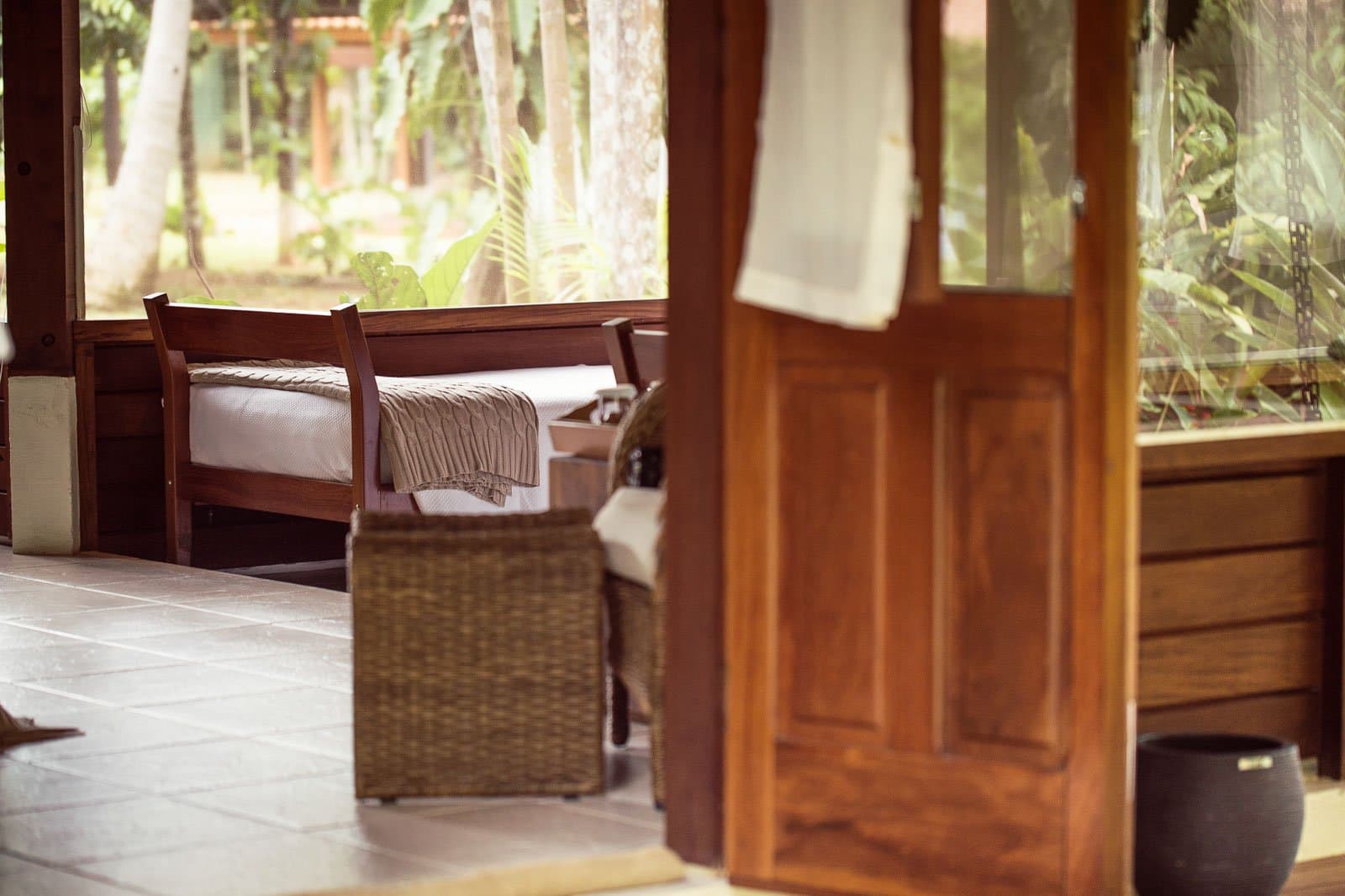 Simple, warm, and quiet, exactly what you need before a 5 AM birding start. Photo: | SamuelMelim Photography Wooden bungalow interior at a birdwatching in the amazon rainforest lodge, showing a single bed with a knitted throw, a wicker armchair, and open screened walls looking out to green forest