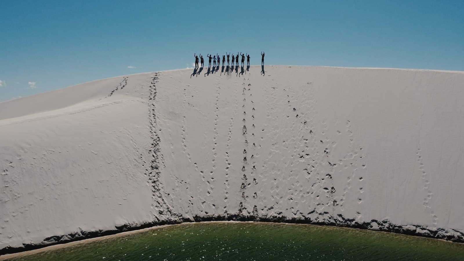 Made it to the top, and the view from here earned every step. A group of travellers silhouetted with arms raised on the crest of a vast white sand dune, their footprints trailing down the slope toward a vivid green lagoon below, on a Lençóis Maranhenses full itinerary